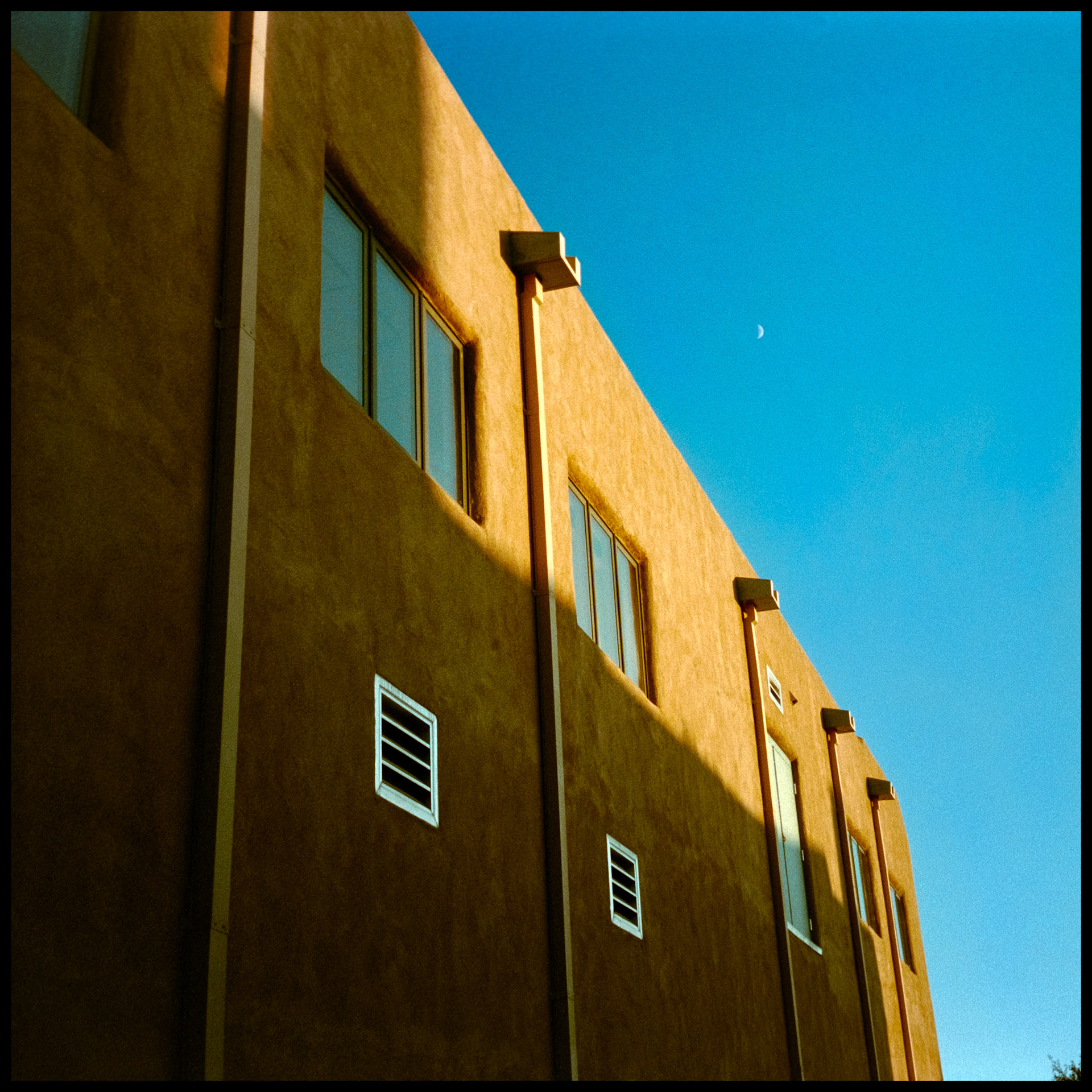 An abstract minimal view of an adobe surfaced wall with long drain pipes partially highlighted by the afternoon sun and the daytime moon in the deep blue sky in the background. Santa Fe, New Mexico USA 1993