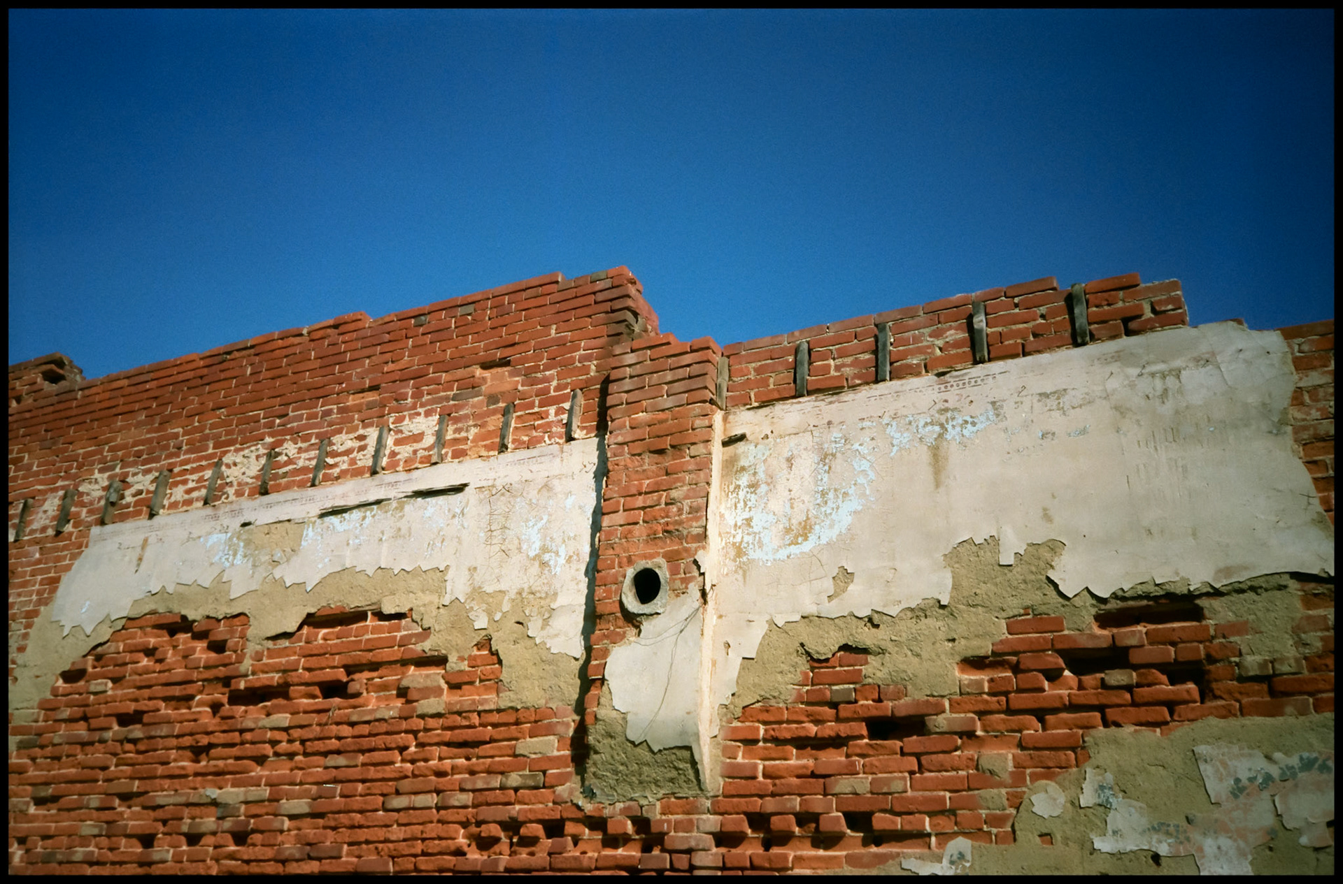 Bare red brick wall of a building during demolition with a heat vent hole as center of attention with a clear blue sky as background. Greencastle, Missouri 1991