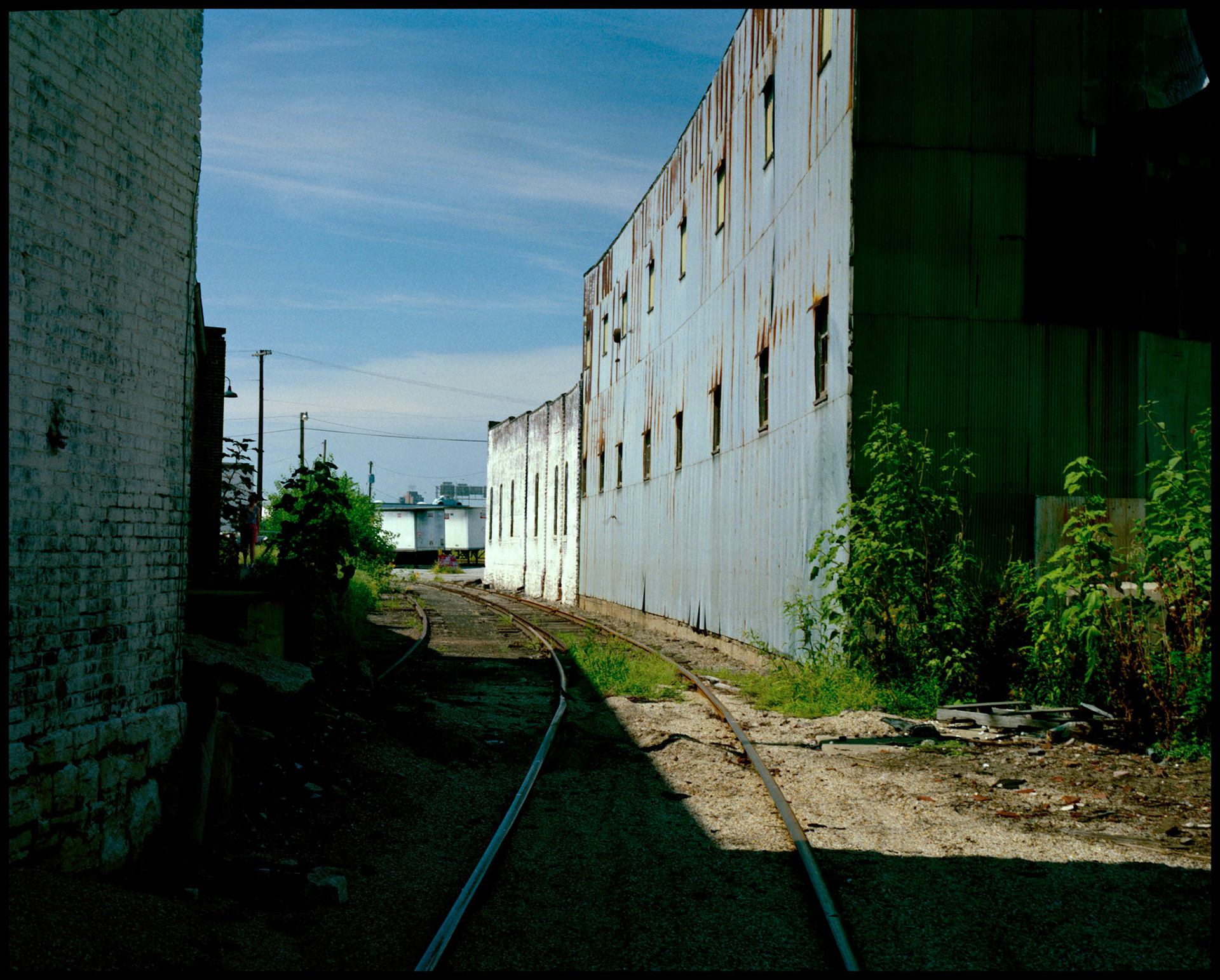 Cityscape of a railroad track curving between two industrial buildings as it emerges from the dark shadow of one building to the late afternoon sunlight. Kansas City, Missouri, 1990