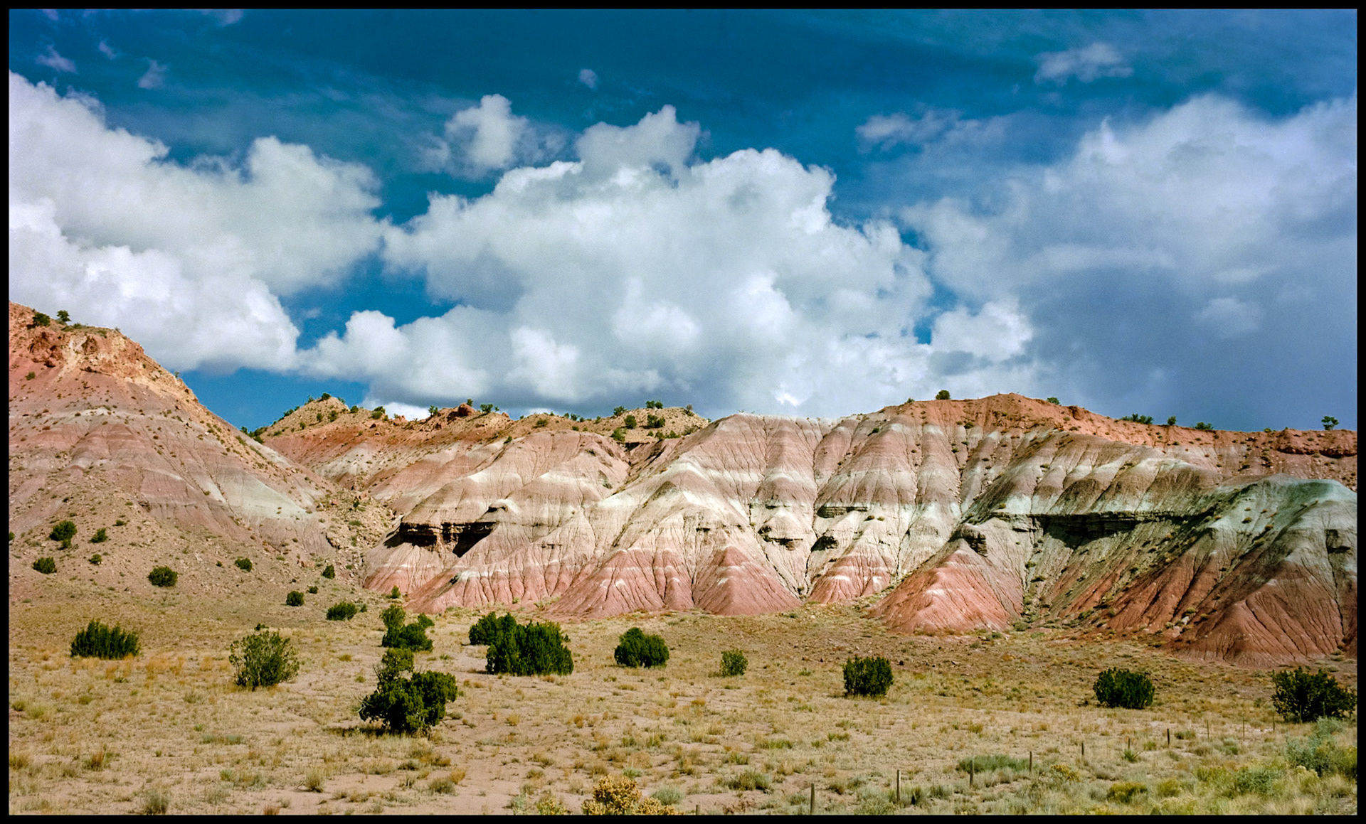 A hillside in New Mexico with muti-colored striped layers and a deep blue sky festooned with billowing white clouds. New Mexico USA 1993