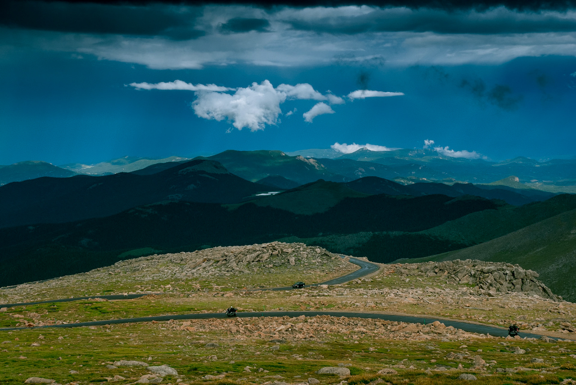 A view from Colorado's Mount Evans Highway, the highest paved road in the USA, showing a car, two motorcycles, and a Bighorn Sheep traveling the road with low hanging clouds against a dark sky above a highlighted mountain top and lake in the background.