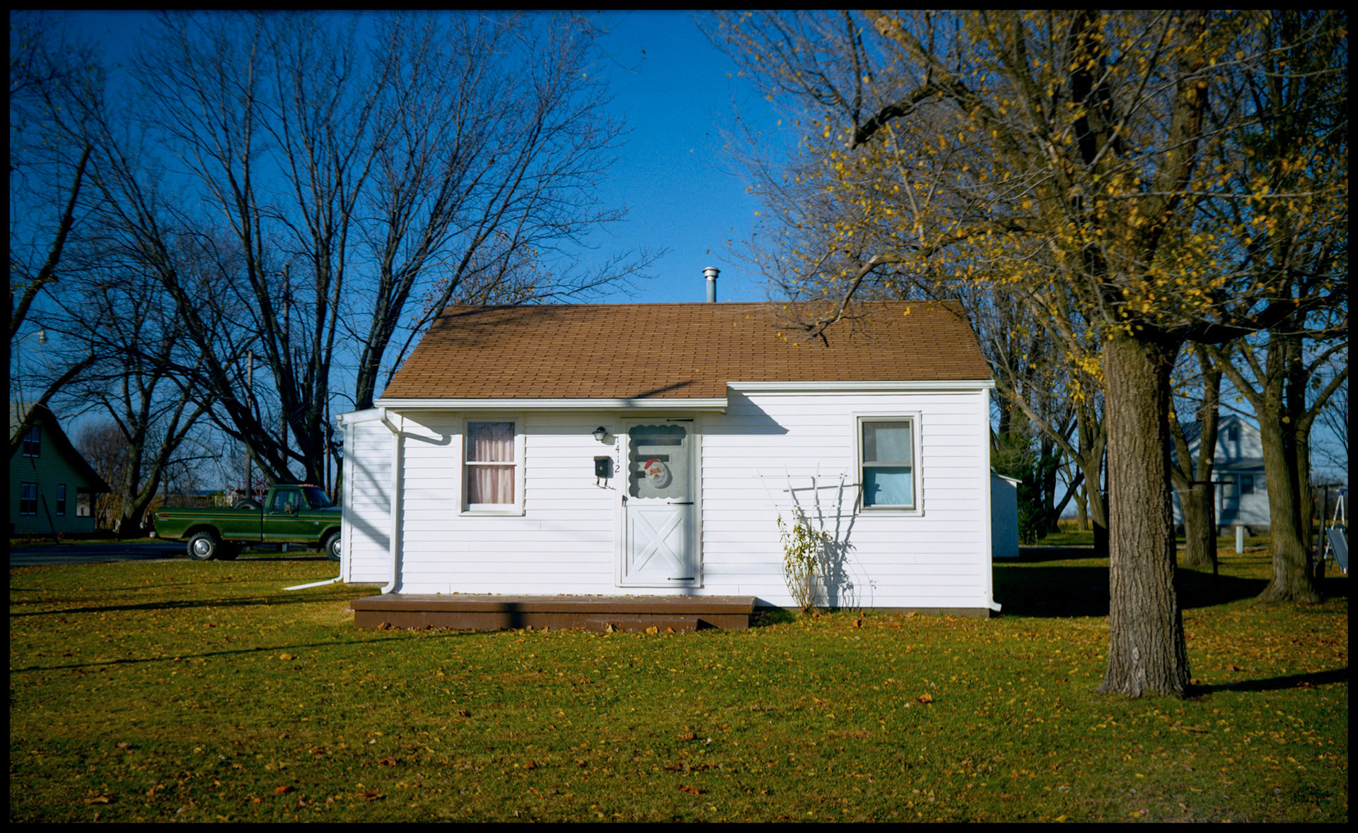 Small white rural house brightly lit by autumn sun with a Santa Claus figure in the front door window.