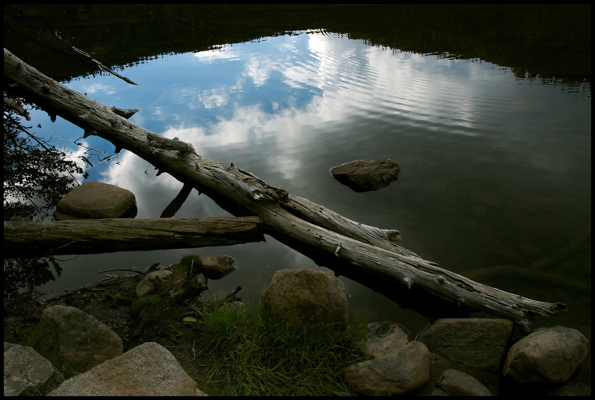 A fallen log in the edge of Bear Lake, Rocky Mountain National Park, Colorado with a rippled reflection of the clouds and blue sky above the surrounding mountain and lakeside boulders in the foreground.  2007
