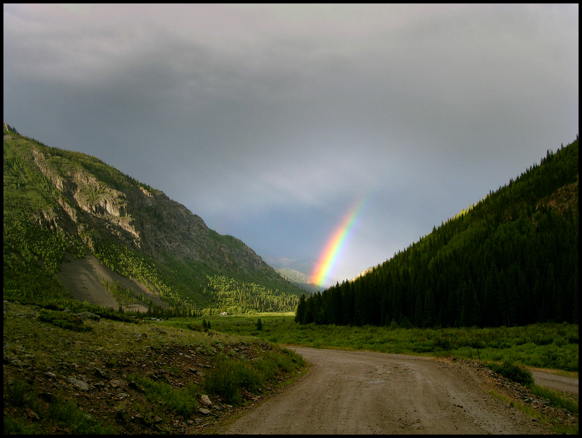 A brightly sunlit rainbow emerging from the mist of a rain shower at the end of a Colorado Rocky Mountain valley between the base of two mountains and a dirt road in the foreground.
