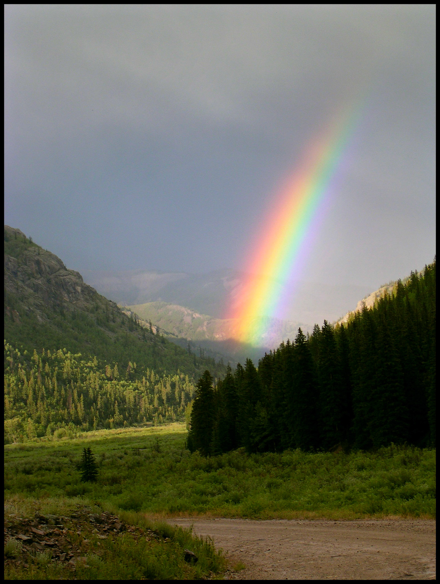 A brightly sunlit rainbow emerging from the mist of a rain shower at the end of a Colorado Rocky Mountain valley between the base of two mountains and a dirt road in the foreground.