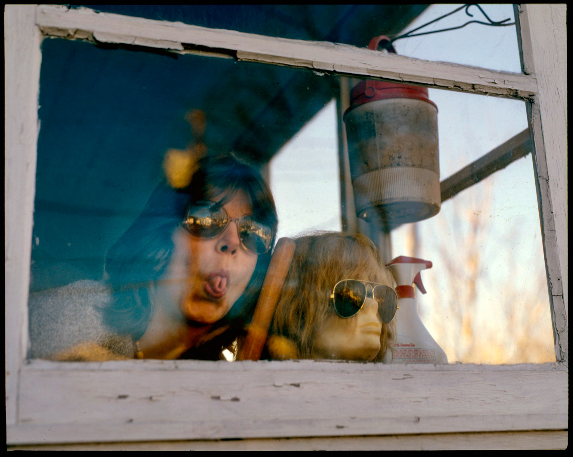 A 1980's whimsical vintage portrait shot through a porch window of a woman posing next to a wigged mannequin head sticking out her rolled tongue. Kirksville, Missouri 1984