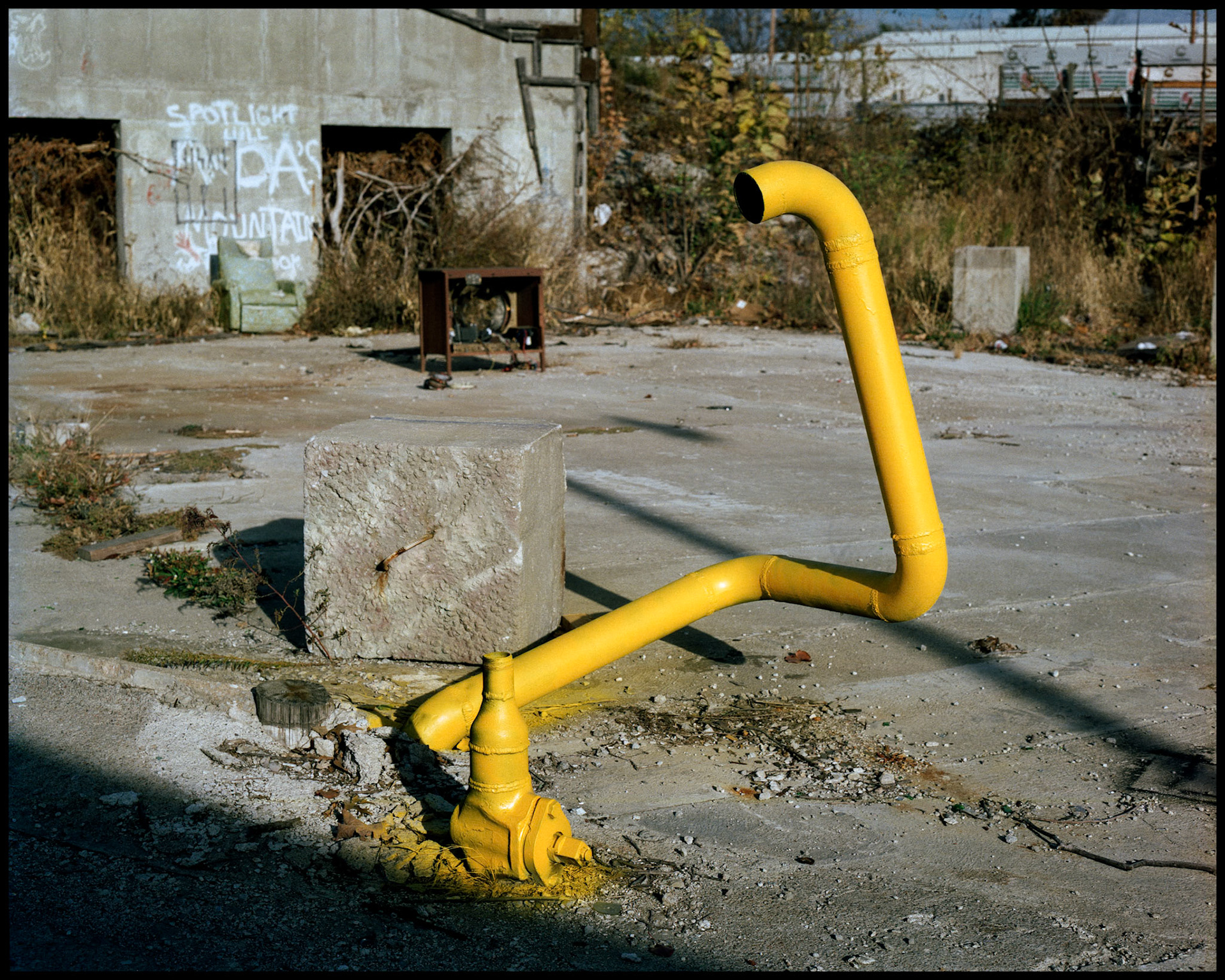 A pipe painted yellow at an abandoned industrial site with a comical scene set up in the background of a junk chair and TV, appearing as if someone had been watching the TV. Columbia, Missouri 1988