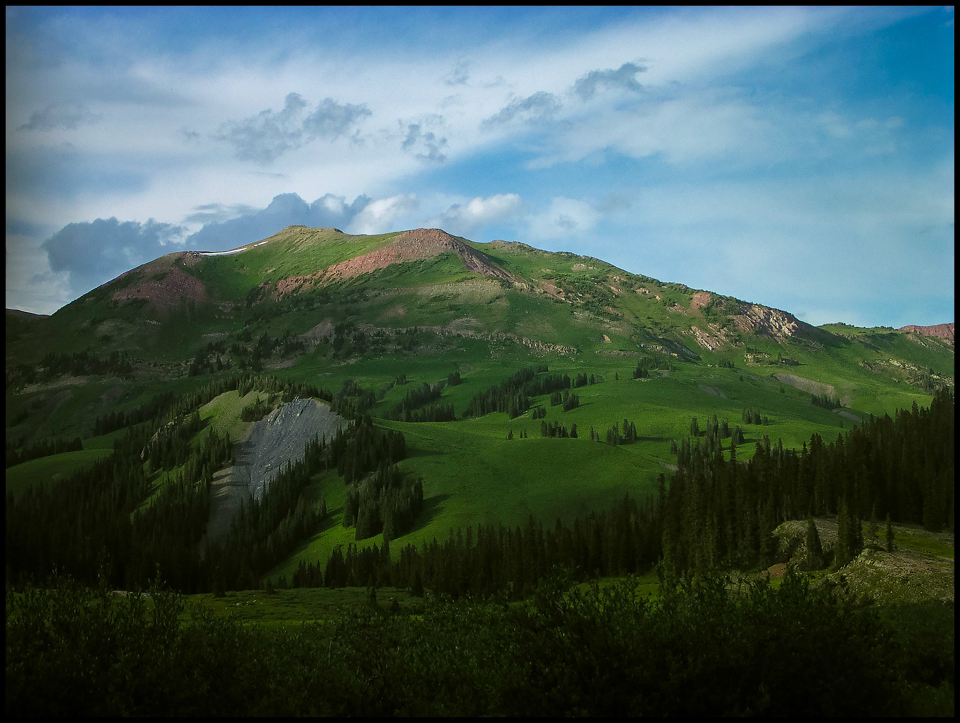 A landscape view of lush green mountain striped with red rock outcroppings and strewn with rows of pine trees with beautiful blue gray clouds in the deep blue sky.