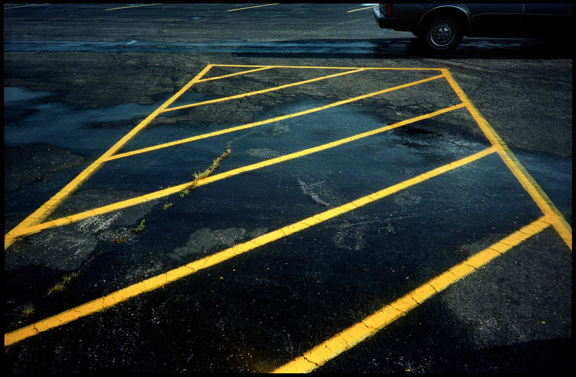 An minimal abstract detail of bright yellow diagonal lines in a black asphalt parking lot making an interesting design with the rear tire of a car in the background. Centralia, Missouri 1994