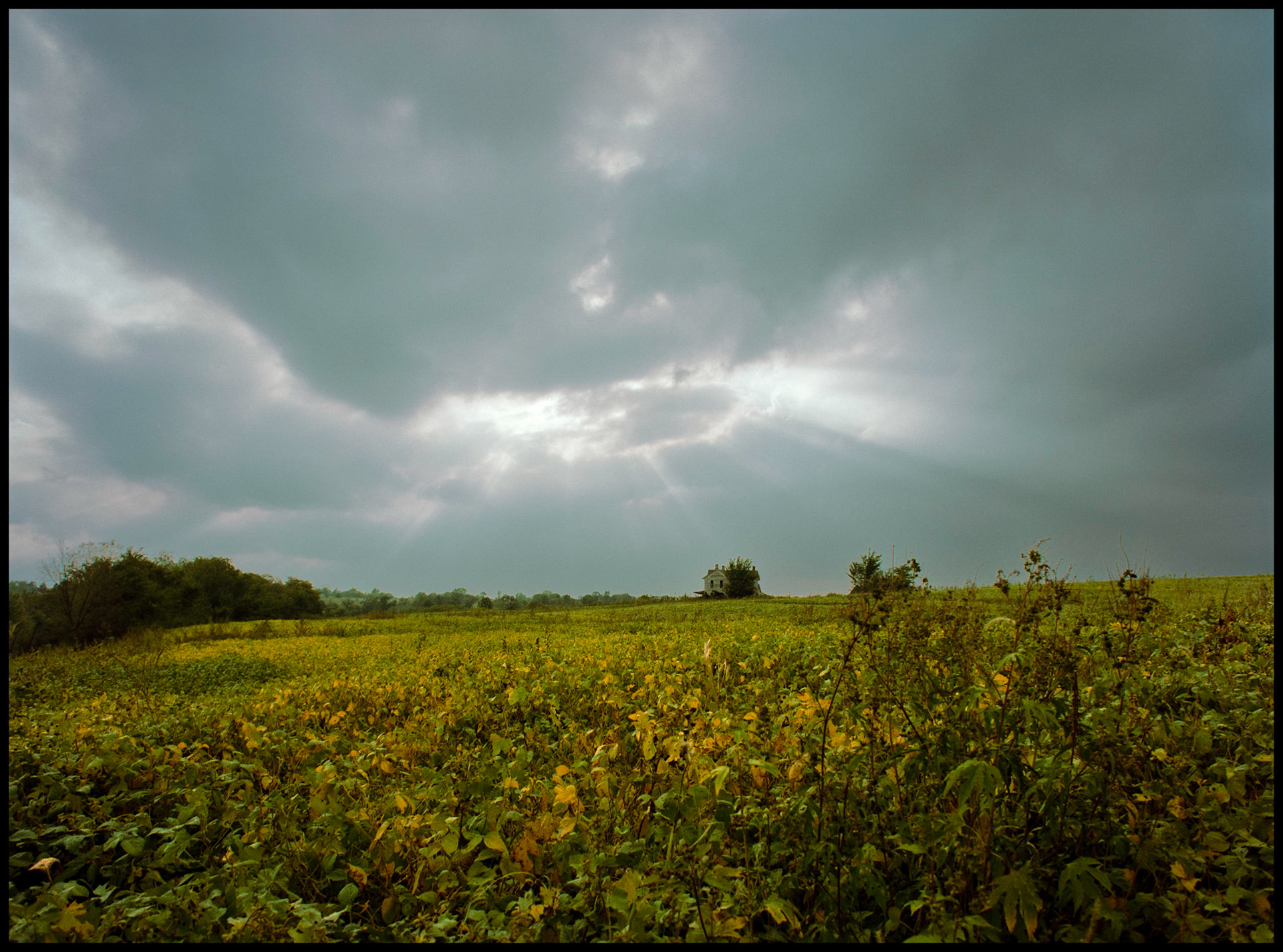 Sun rays beaming through an opening in dark clouds above a colorful pasture and abandoned farm house. Missouri, USA, 1984