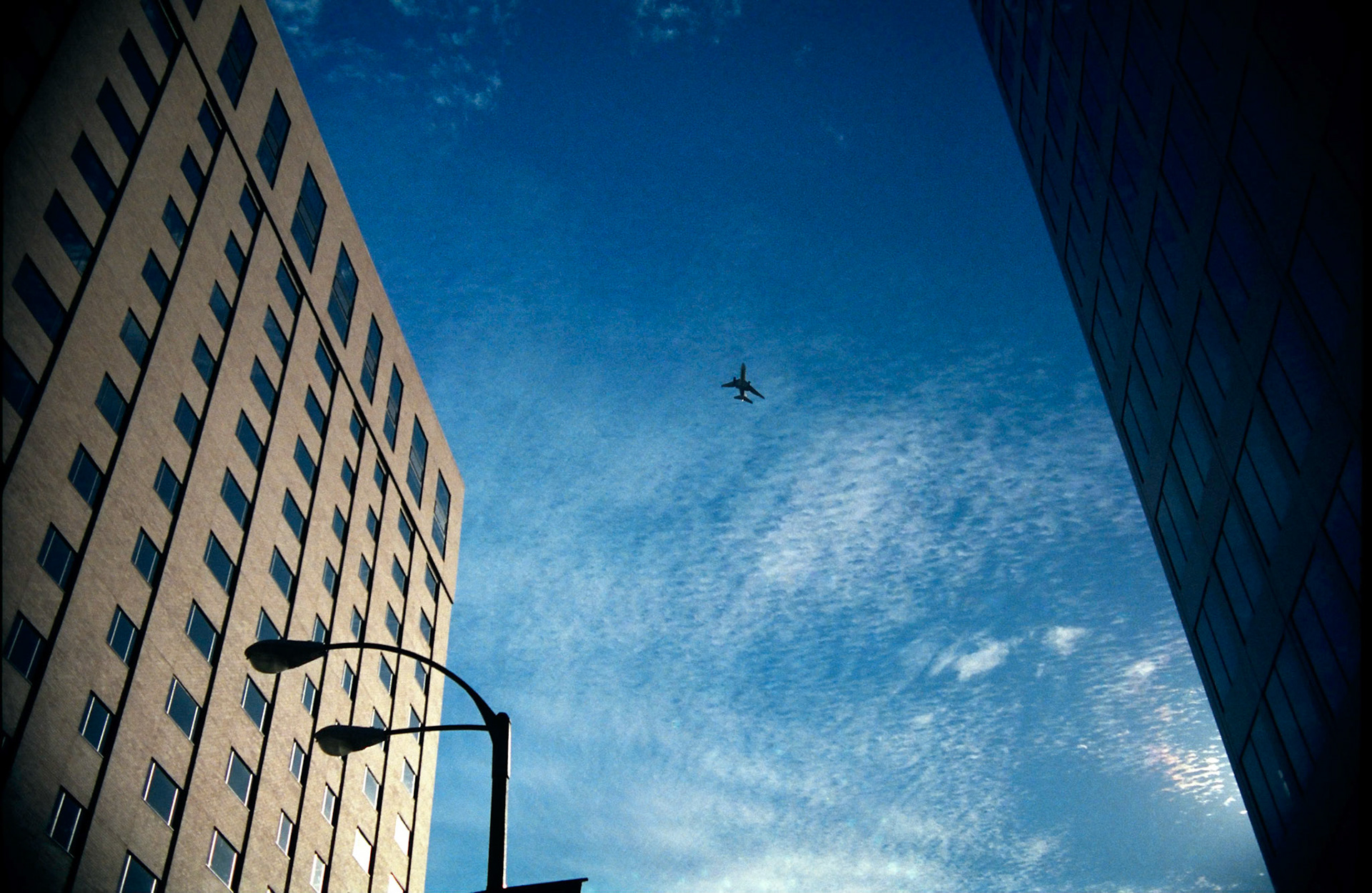 A minimal urbanscape with an approaching airliner framed between two looming builidings and silhouetted streetlamp against a rich deep blue sky with a few textured cirrus clouds. Part of a series shot one warm afternoon in November, 1988 called An Afternoon in St. Louis (a subset of my Industrial Geometry series).