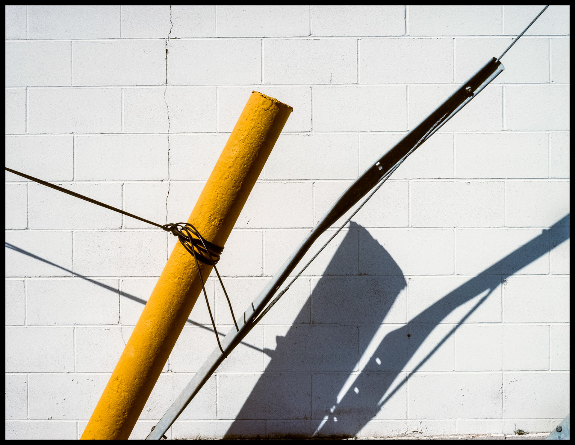 Leaning yellow parking lot safety bollard with cable tied to it seemingly supporting it along with a guy wire and bent cover and their shadows against a white brick wall. Kirksville Missouri 1985