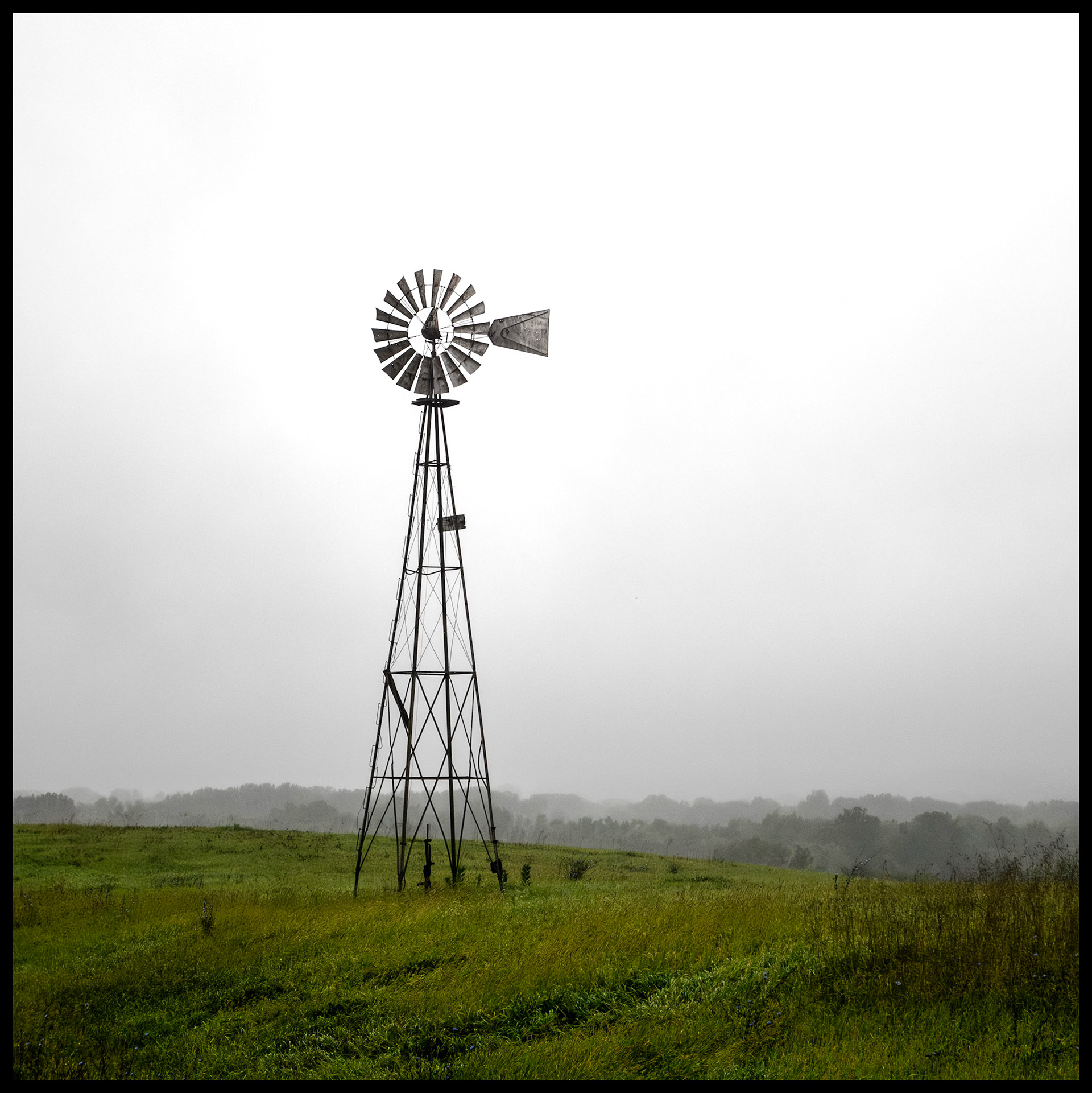 A solitary windmill at the edge of a green field silhoutted against the overcast sky on a rainy and foggy September morning. Located on Indian Hill near South Gifford, Missouri, 2023.