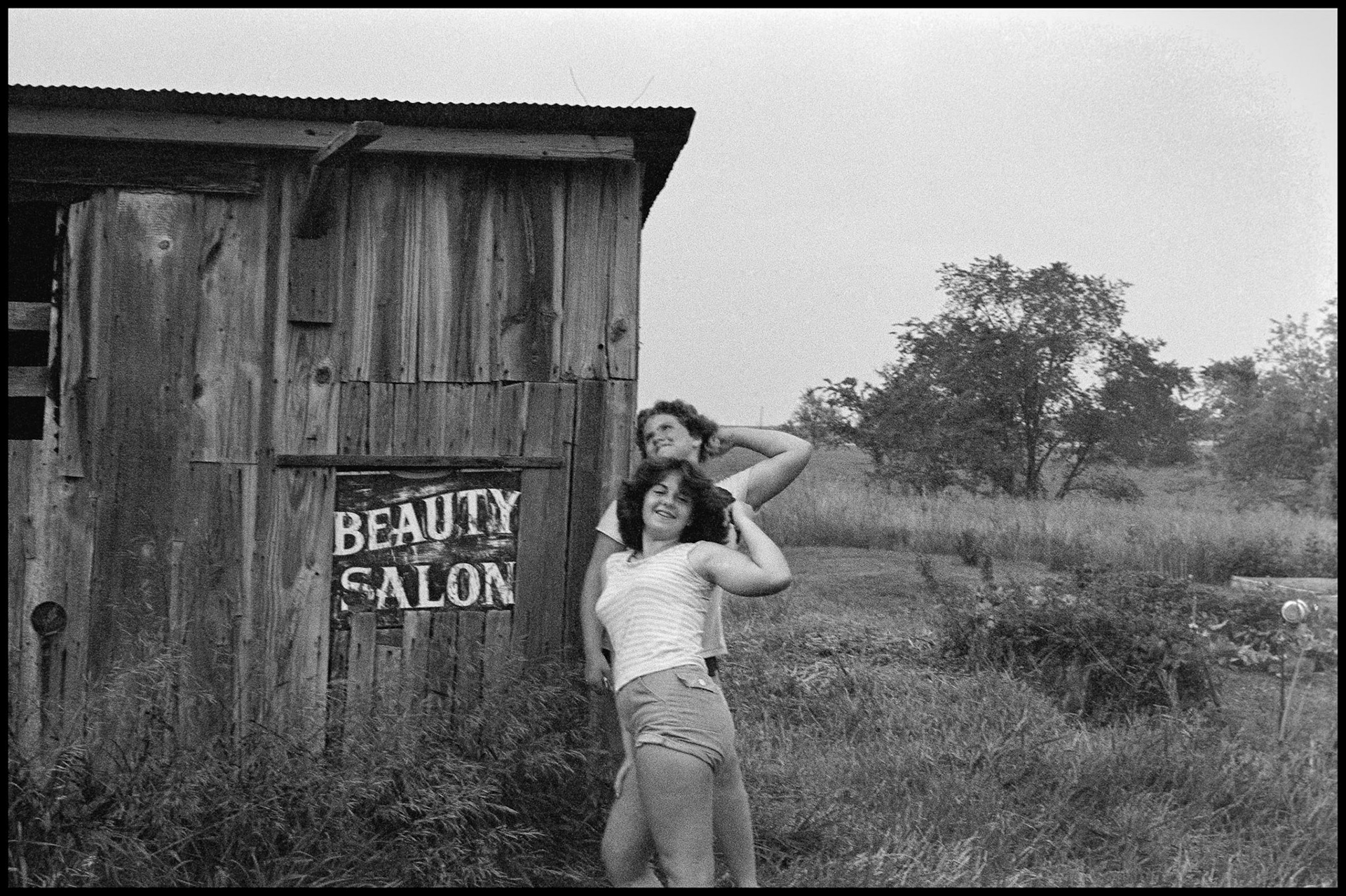 A 1970's vintage image of two teenage sisters posing next to an weathered shed that has a beauty salon sign patching an old window. Green City, Missouri 1979