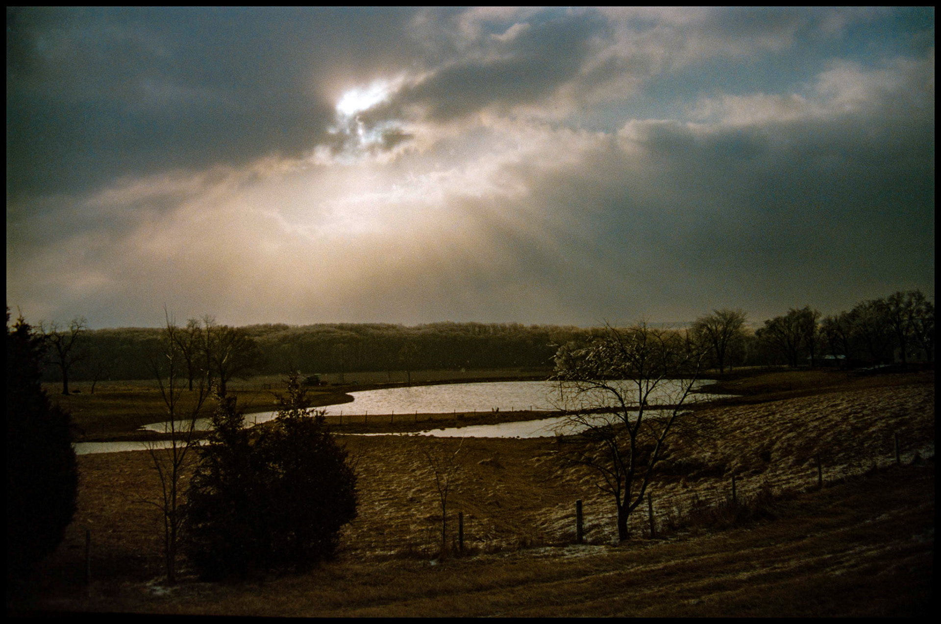 The sun breaking through some dark clouds on a frosty winter morning over a farm pond in central Missouri. 1989