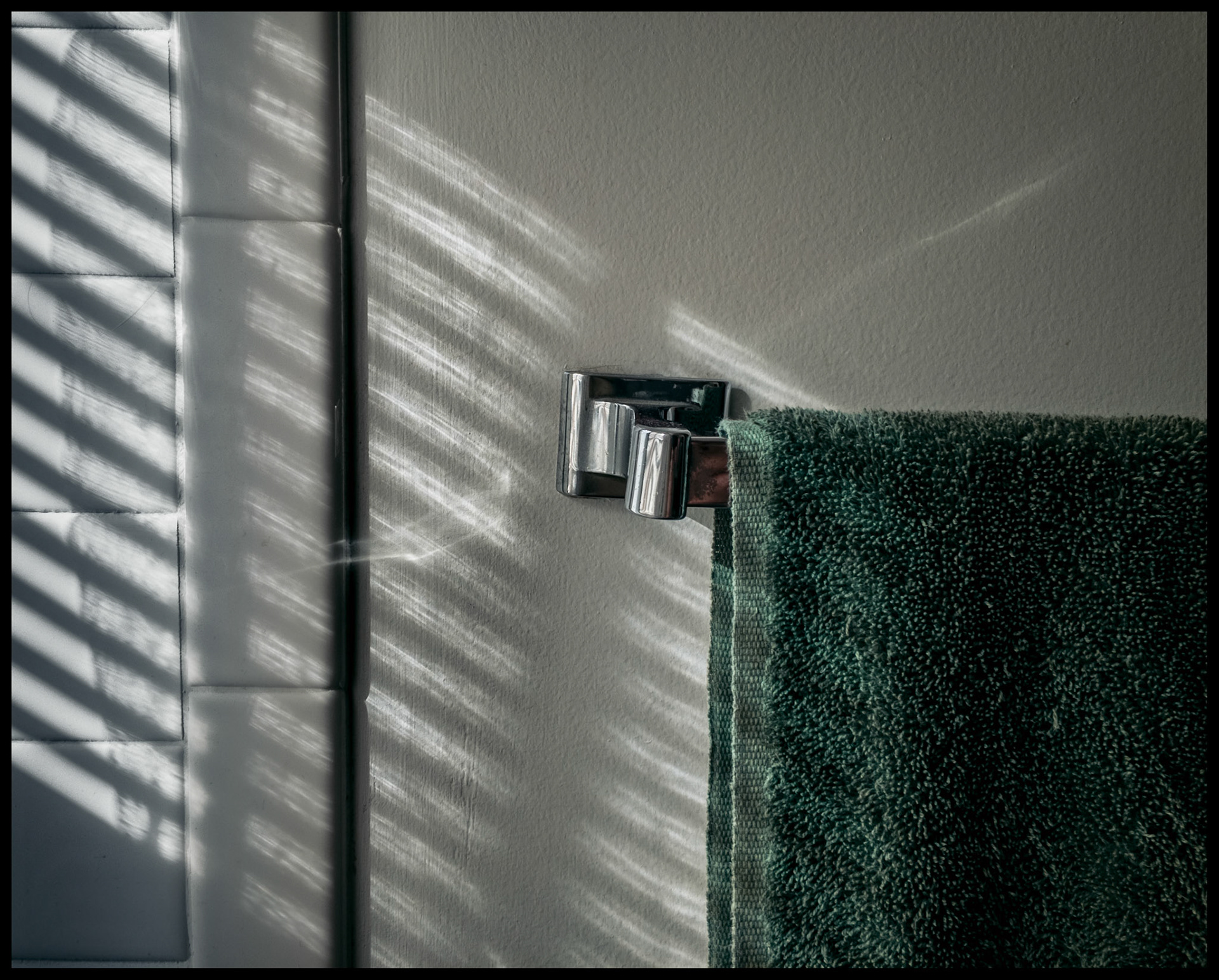 A minimal abstract still life view of highlights and shadows from a window blind on a bathroom wall and towel rack. Kirksville, Missouri 2022