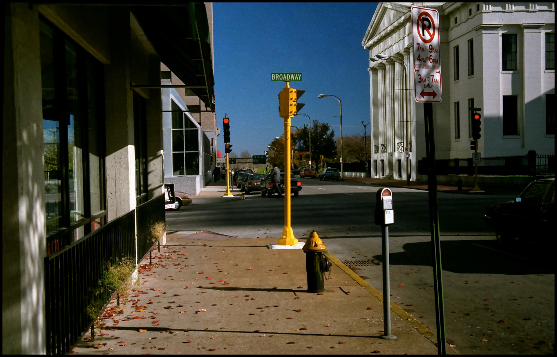 A vintage urban street scene of downtown St. Louis Missouri in 1988 featuring a brightly sunlit yellow stoplight surrounded by a fire hydrant, parking meter, and various street signs. 1988