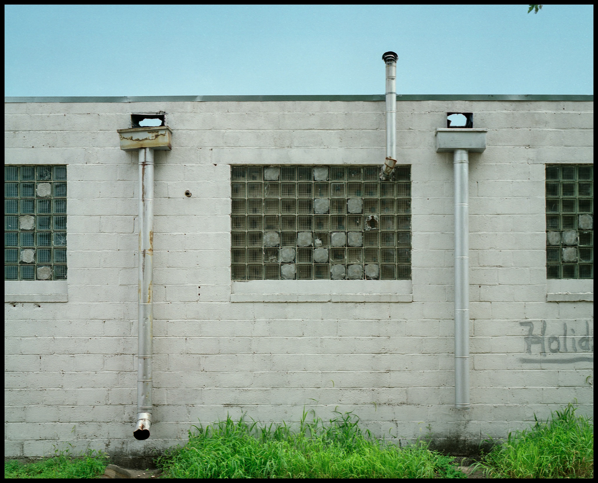 An urban detail image of drain and vent pipes on the side of a white cement block building with glass block windows and green grass in the foreground in an industrial area of Columbia, Missouri in 1988