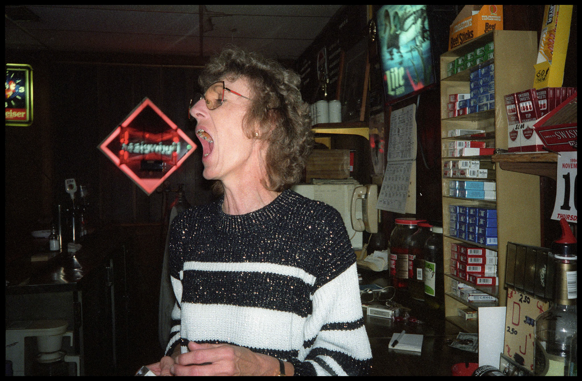 A whimsical photo of a bartender in a small town tavern yelling out to somebody above the music with an hilarious alignment of a Budweiser sign in the background. Greencastle, Missouri, 1993