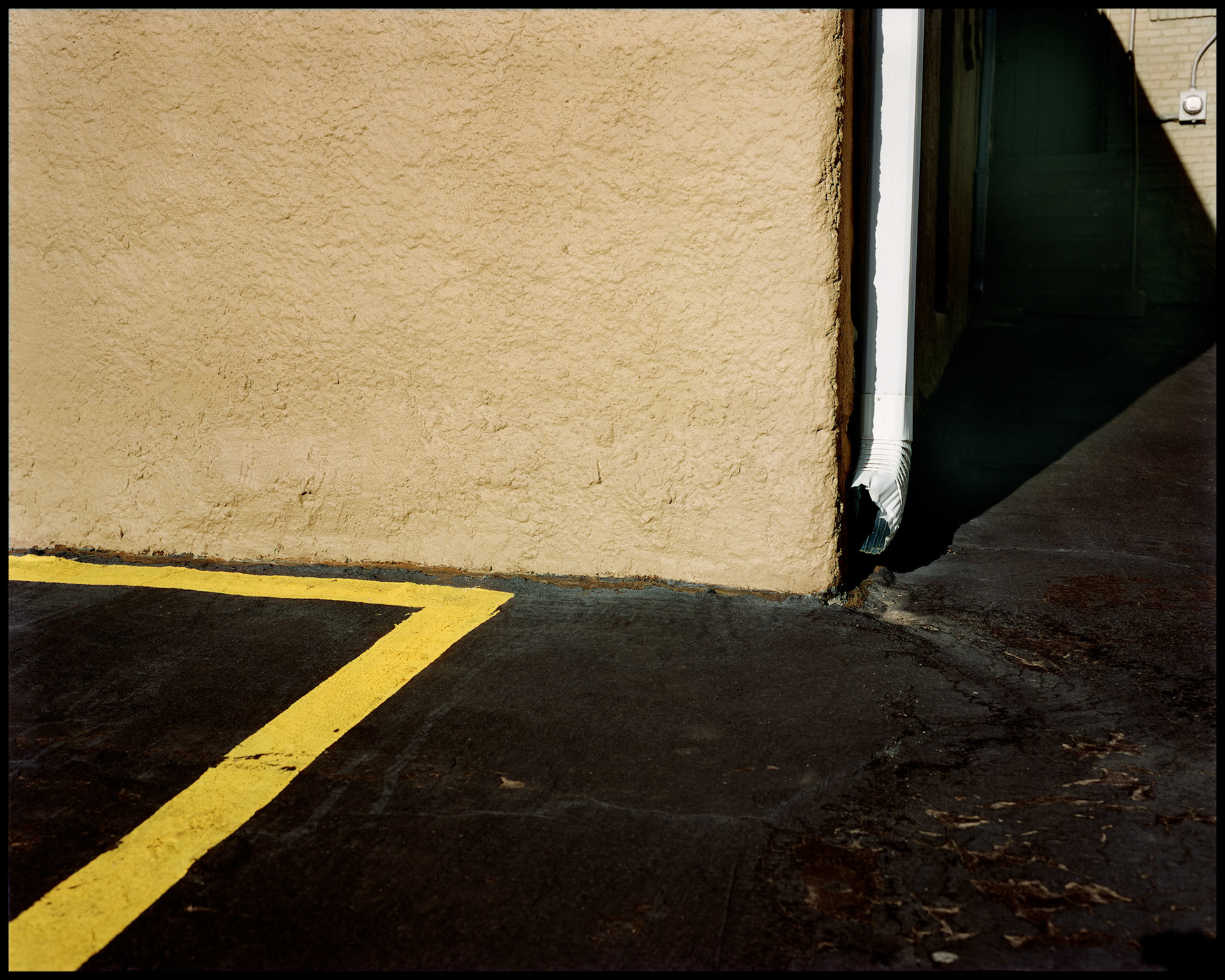 A minimal abstract urban detail of a parking lot using a wall, downspout, parking line, and an electric meter as design elements. Baker Gallery, 45th and State Line Road, Kansas City Kansas 1984