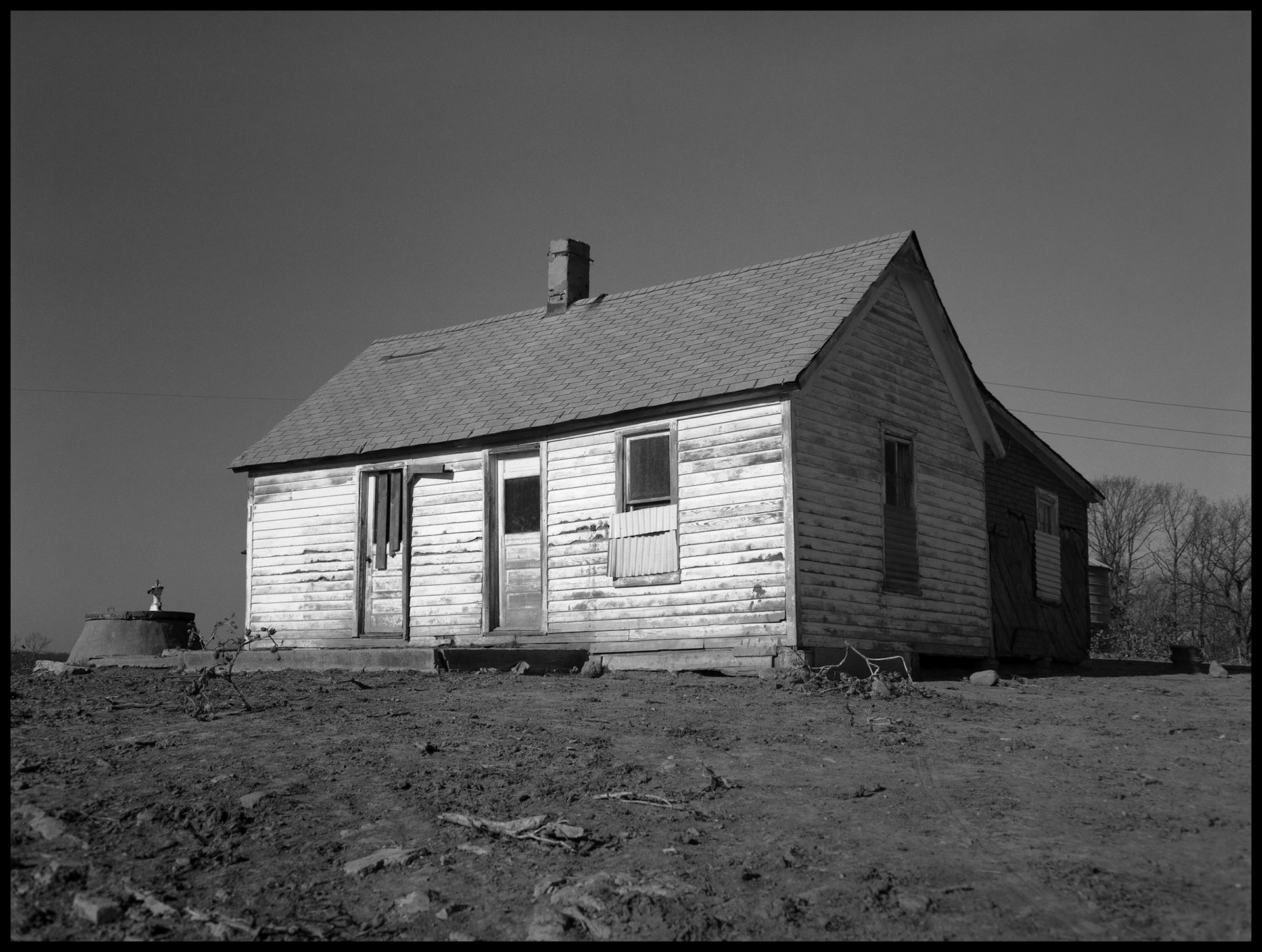 A vintage black and white view of a boarded-up abandoned farmhouse and well pump highlighted by the winter afternoon sun. Near Nind, Missouri 1979