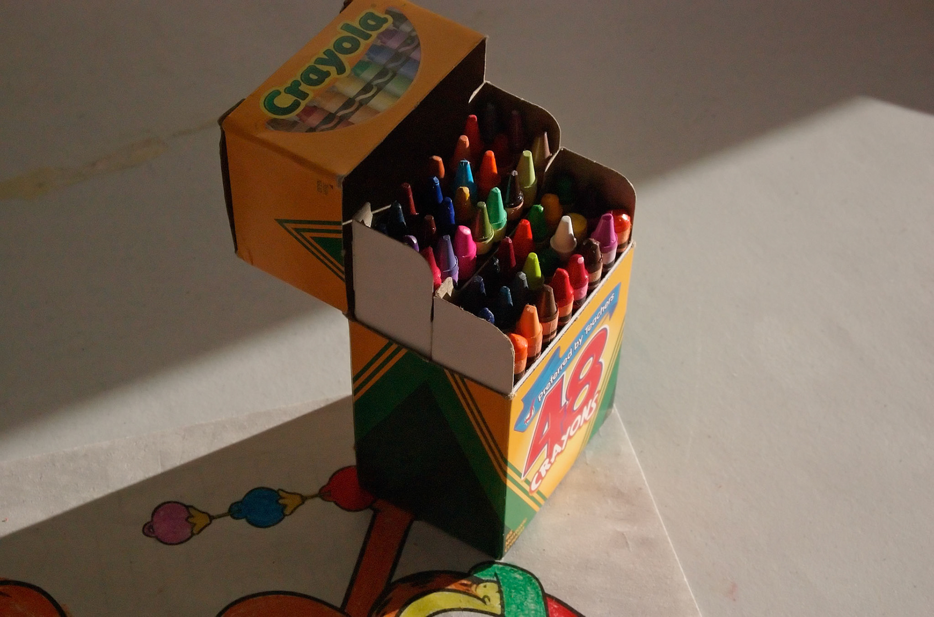 An open box of used crayons on top of a "colored" page, sidelit by the sun.