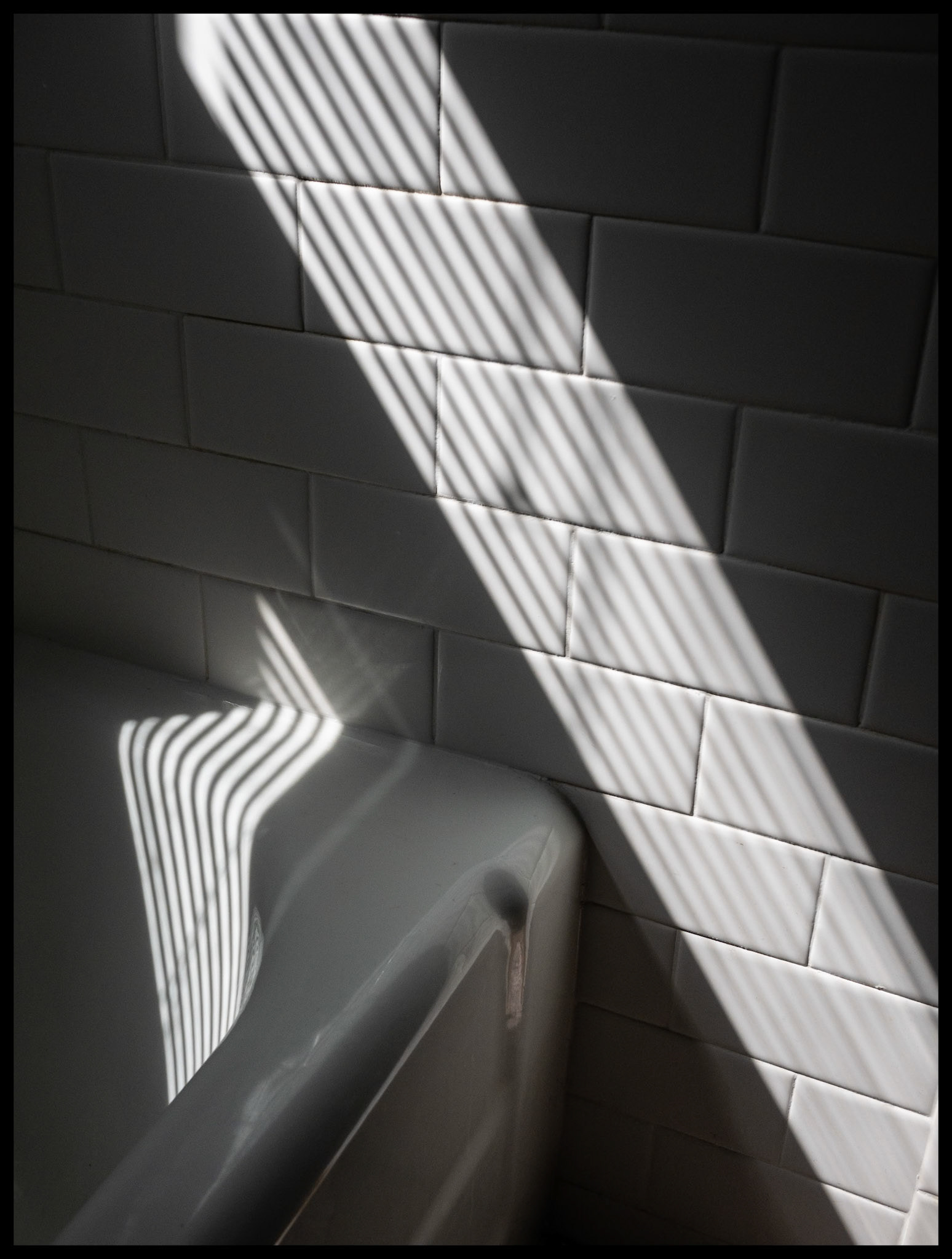 An abstract detail of the highlights and shadows of the sun shining through a window blind onto a bathtub and tile wall creating many interesting lines. Kirksville, Missouri 2024