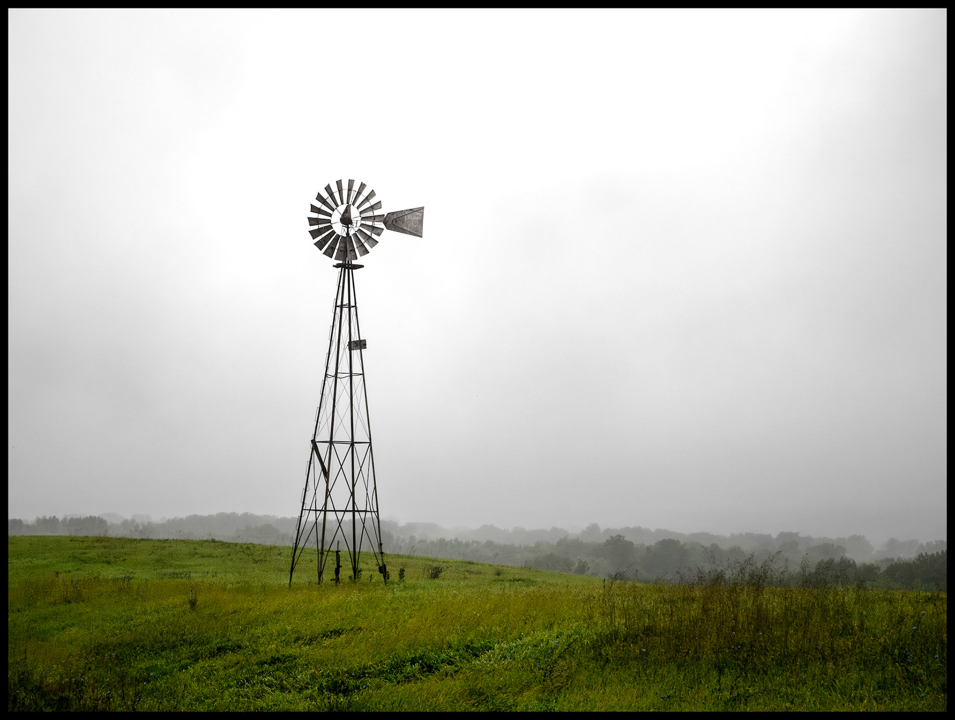 A solitary windmill at the edge of a green field silhoutted against the overcast sky on a rainy and foggy September morning. Located on Indian Hill near South Gifford, Missouri, 2023.