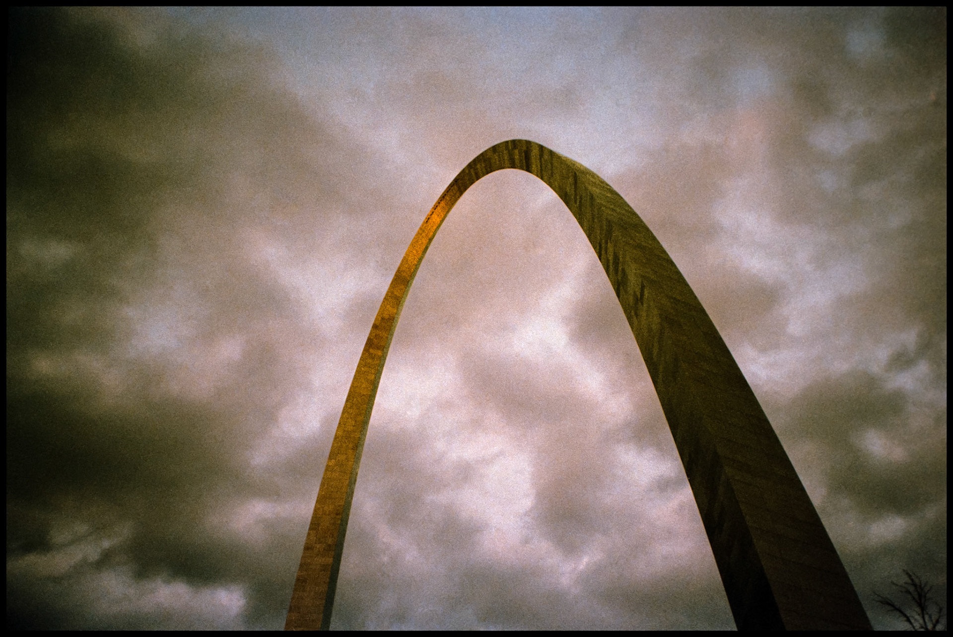 A grainy upward view of the Gateway Arch in St. Louis, Missouri at dusk with a golden highlight from the last rays of the setting sun. 1986
