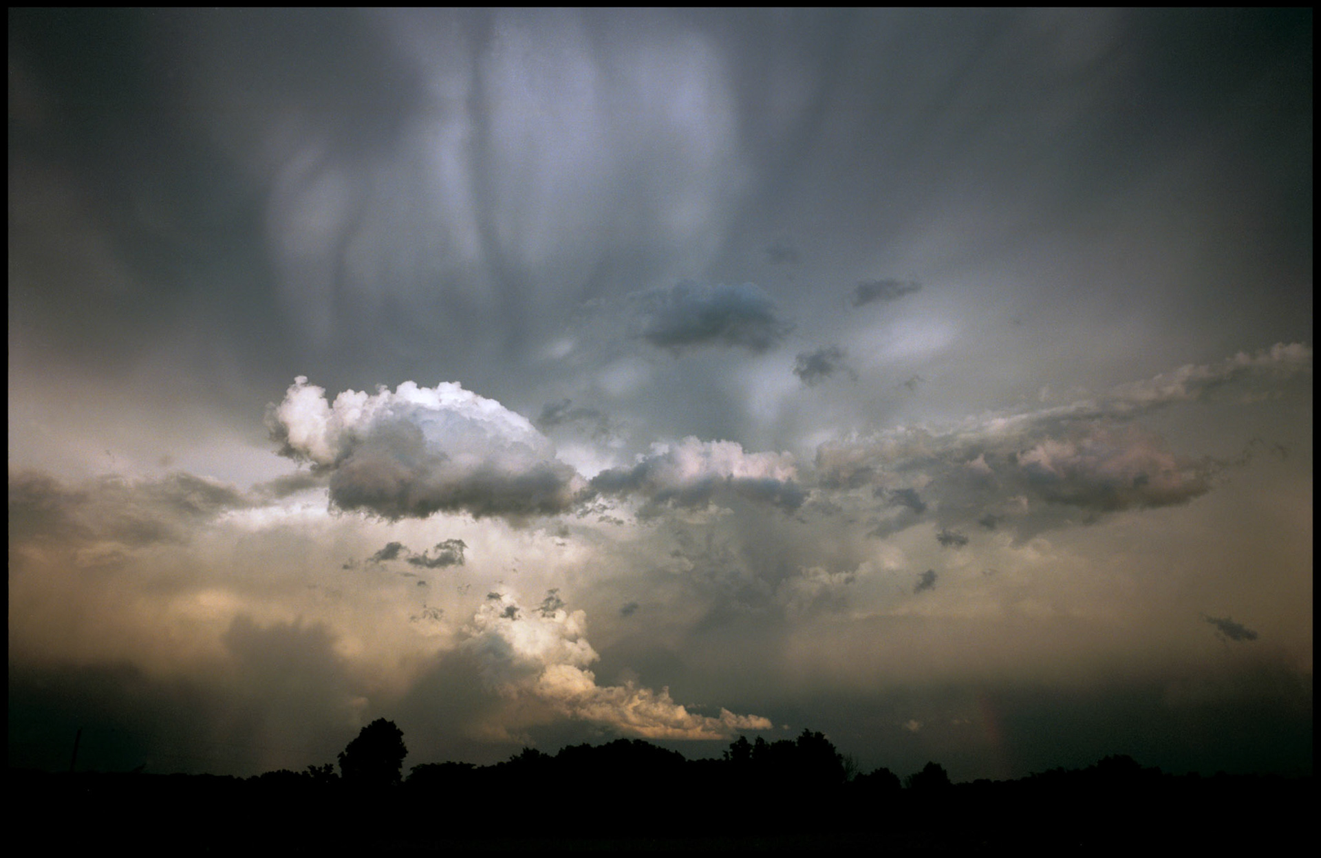 Dramatic turbulent clouds of an approaching storm front brightly lit from above by the evening sun through an opening in the clouds with silhouetted trees in the foreground and a hint of a rainbow on the horizon. My front yard near Renick, Missouri, 1996.