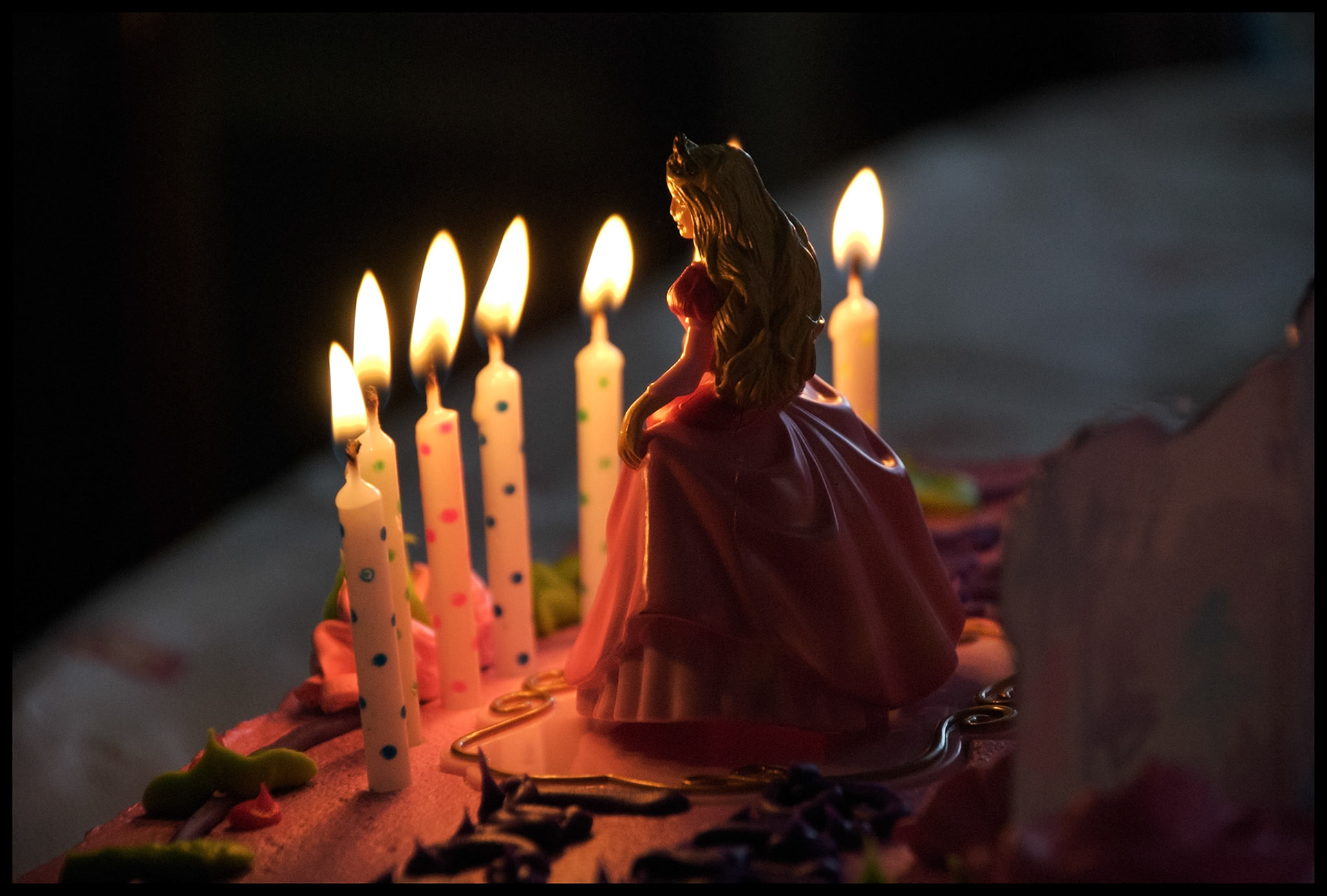 A closeup still life of a childs birthday cake adorning a princess figurine, candlelit by the seven polka-dot birthday candles resting on pink icing. Renick, Missouri 2007