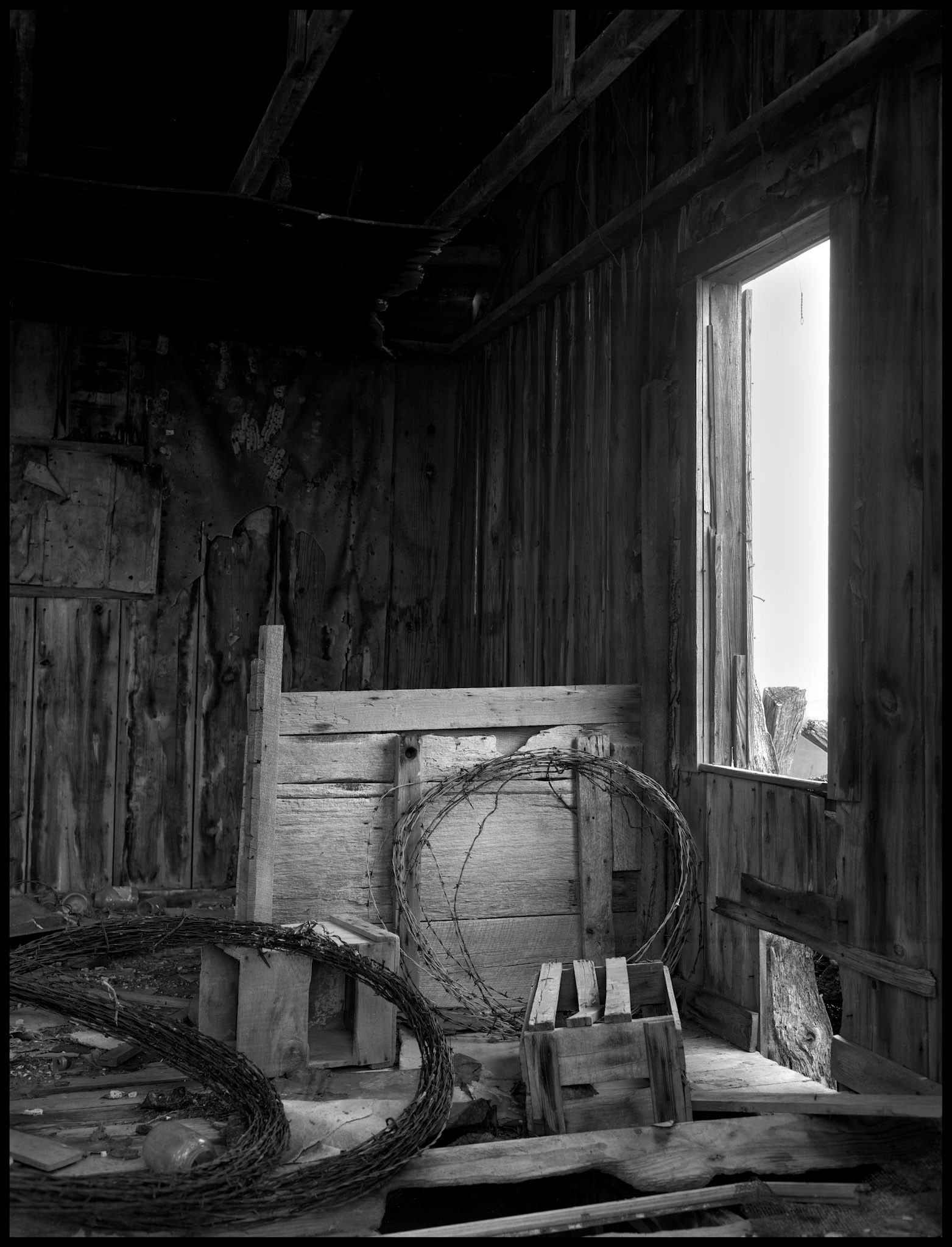 A rustic black and white still life of the interior of an abandoned farm building with spools of barbed wire propped against the former stall wall. Near New Boston, Missouri, 1980