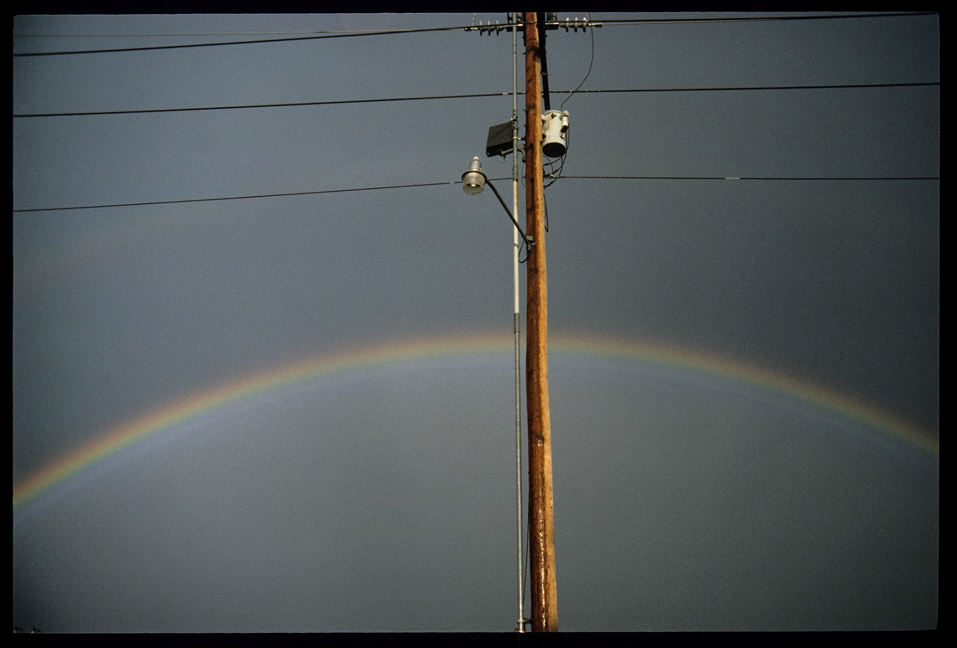 Rainbow behind highlighted power pole with streetlight against a dark grey sky. Kirksville, Missouri 1986