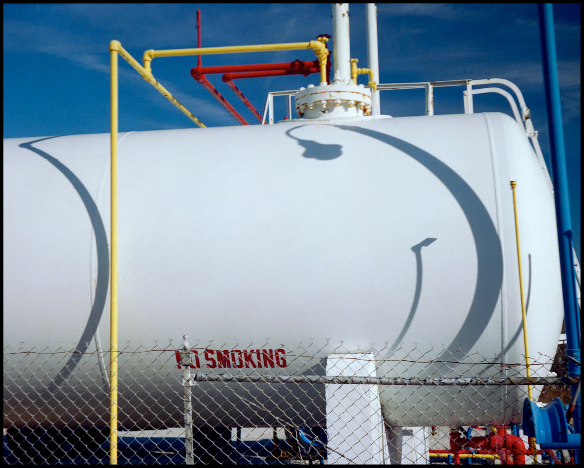 The curved shadows of pipes and a lightpole on a brilliant white propane gas tank adorned with colorful pipes. Columbia, Missouri 1989
