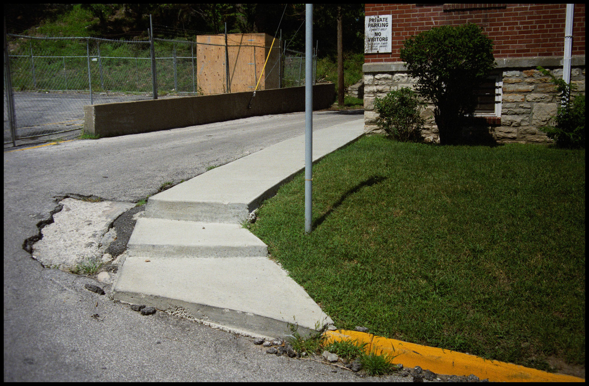 A minimal abstract detail view of a curved sidewalk, flagpole, and curb next to a brick building. Kansas City, Missouri 1990