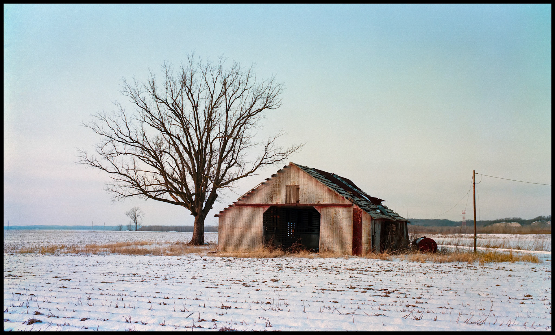 A metal barn basked in the warm pastel light of the dusk sun in a snow covered plowed field next to a large bare tree. Missouri River bottoms near Wooldridge, Missouri a few months before the great flood of 1993.