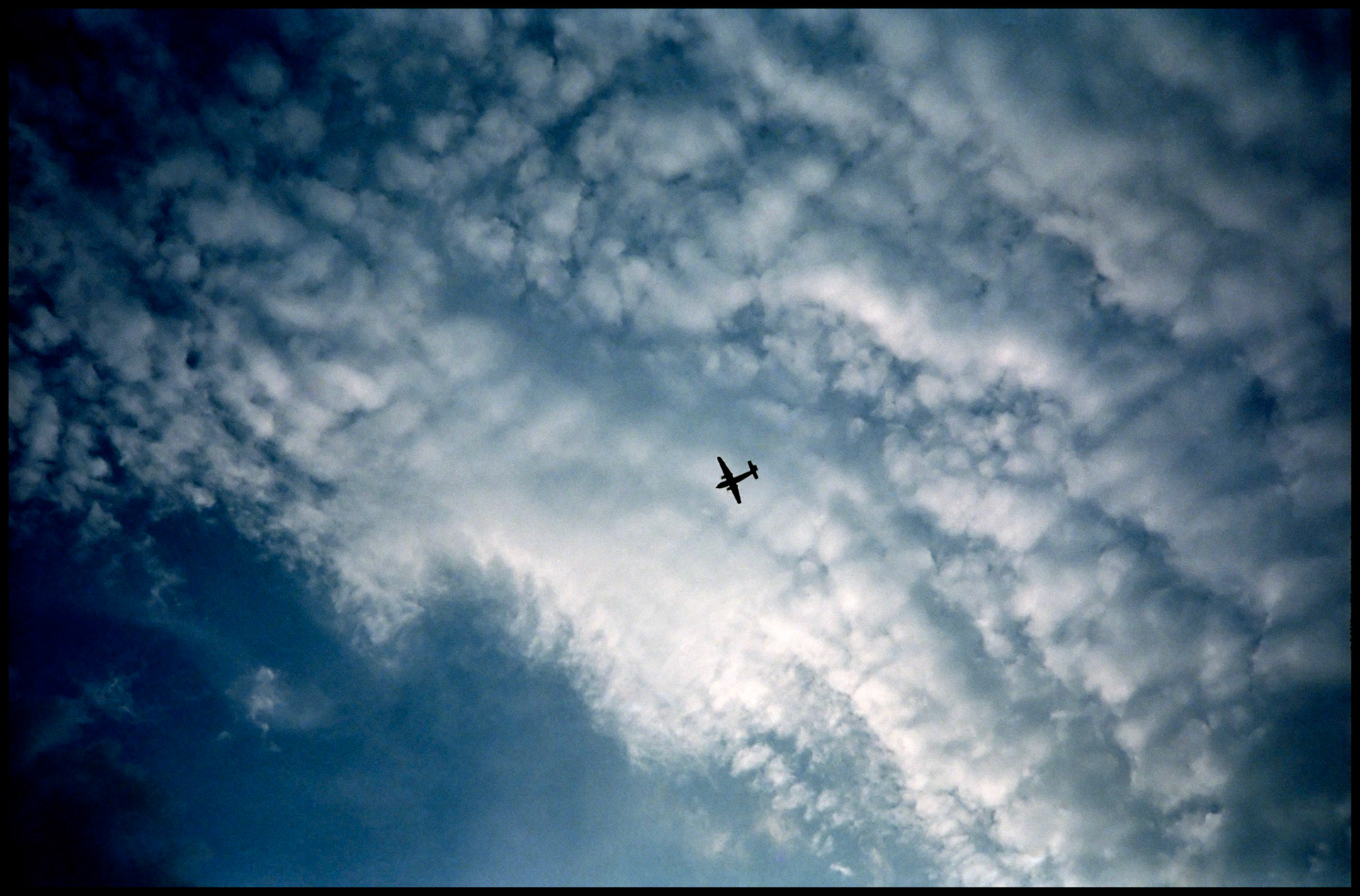 A dreamy view of a silhouetted antique airplane from below against sidelit shimmering white puffy clouds and a deep blue sky. Near Hinton, Missouri USA 1991