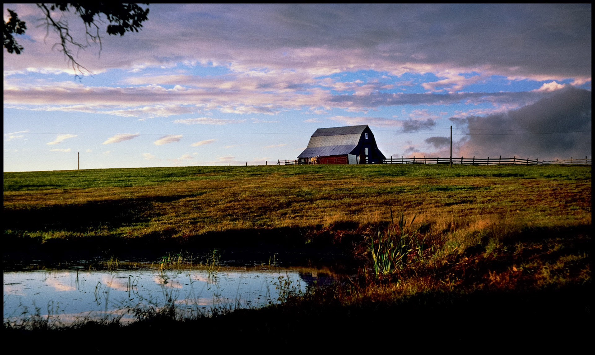 An idyllic scene of a horse in front of a barn at the edge of a pasture with purplish blue/gray clouds in the background and a pond reflecting the clouds in the foreground. Near Hinton, Missouri 1990