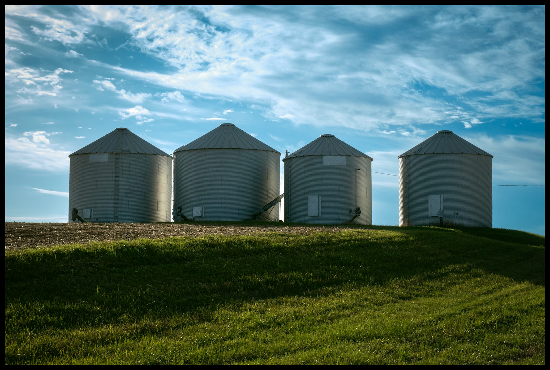 Four backlit grain bins on a hill at the edge of a soybean field right after harvest framed by a blue sky with whispy clouds. Near Ethel, Missouri. 2023