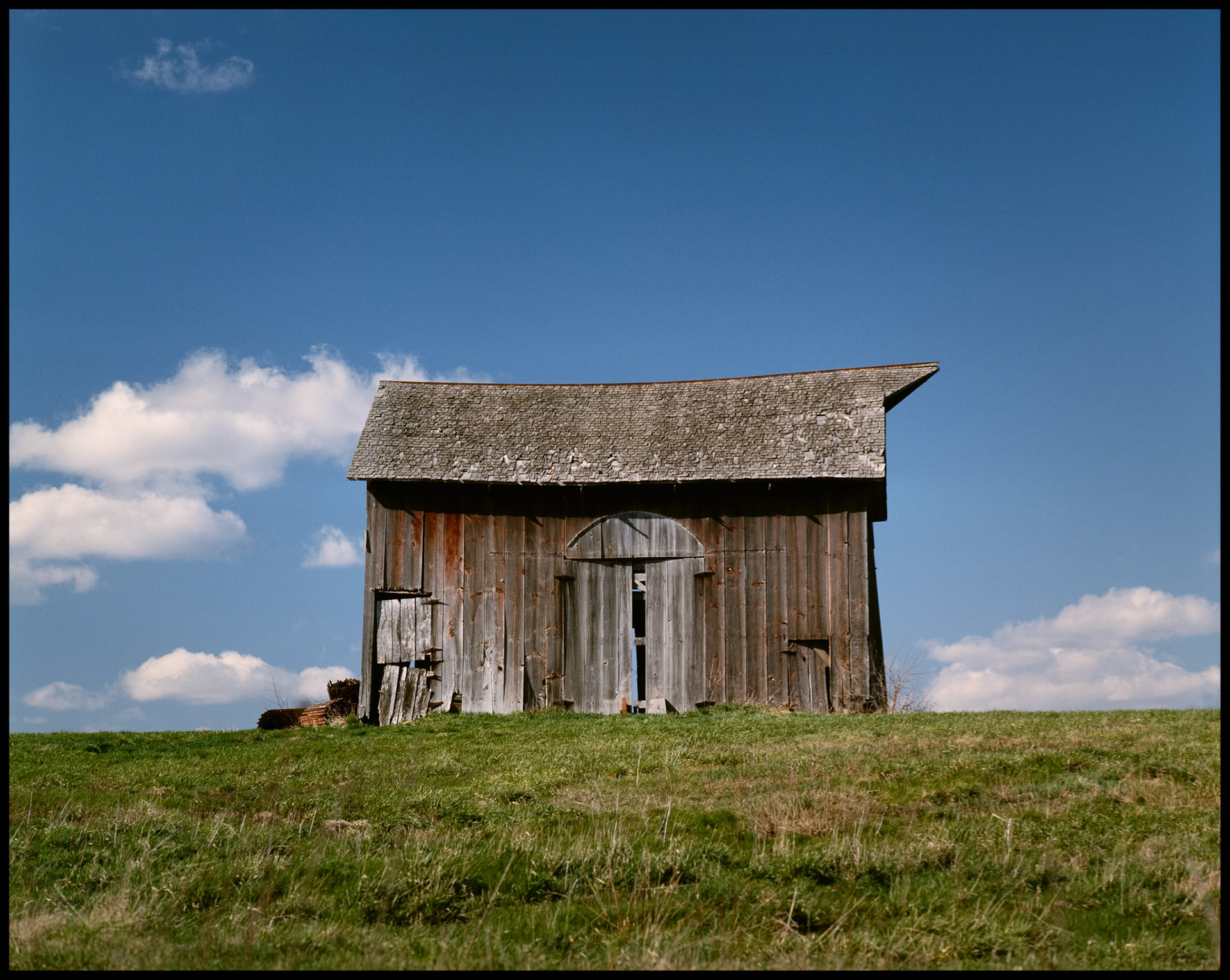 An old barn that was, at one time, red on a hillside in Adair County in Northeast Missouri with puffy clouds dotting the blue sky background. 1985