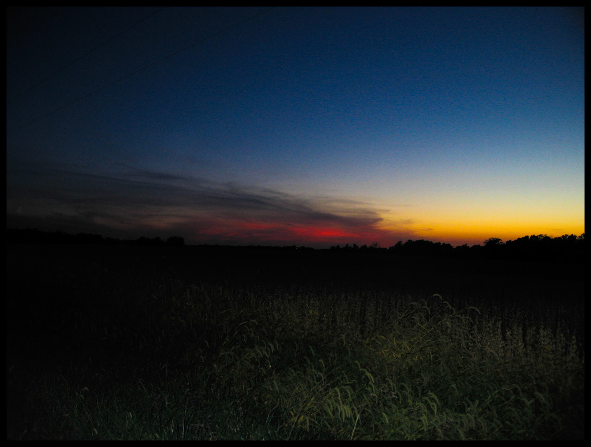 The horizon with the last peek of red sunset shining through an eyelike gap in the whispy clouds. Contrasted by the yellow orange cloudless sky and the deep blue of the darkening sky with hints of the field grass in the foreground. Near Moberly Missouri. 2006