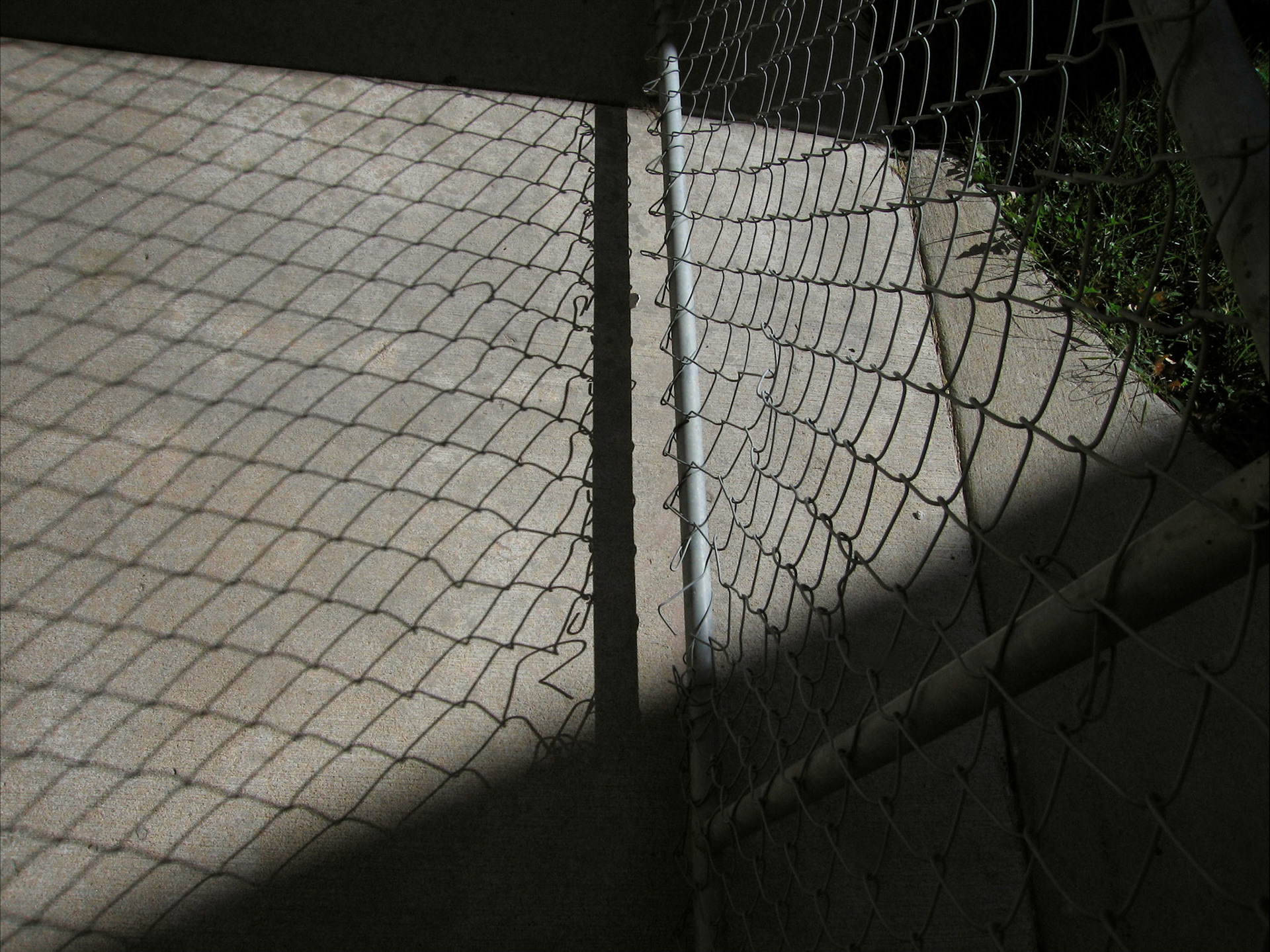 An minimal abstract detail of the bottom rail of a chain link fence higlighted by the afternoon sun while casting deep patterned shadows on the sidewalk below. Truman State University campus, Kirksville, Missouri 2006