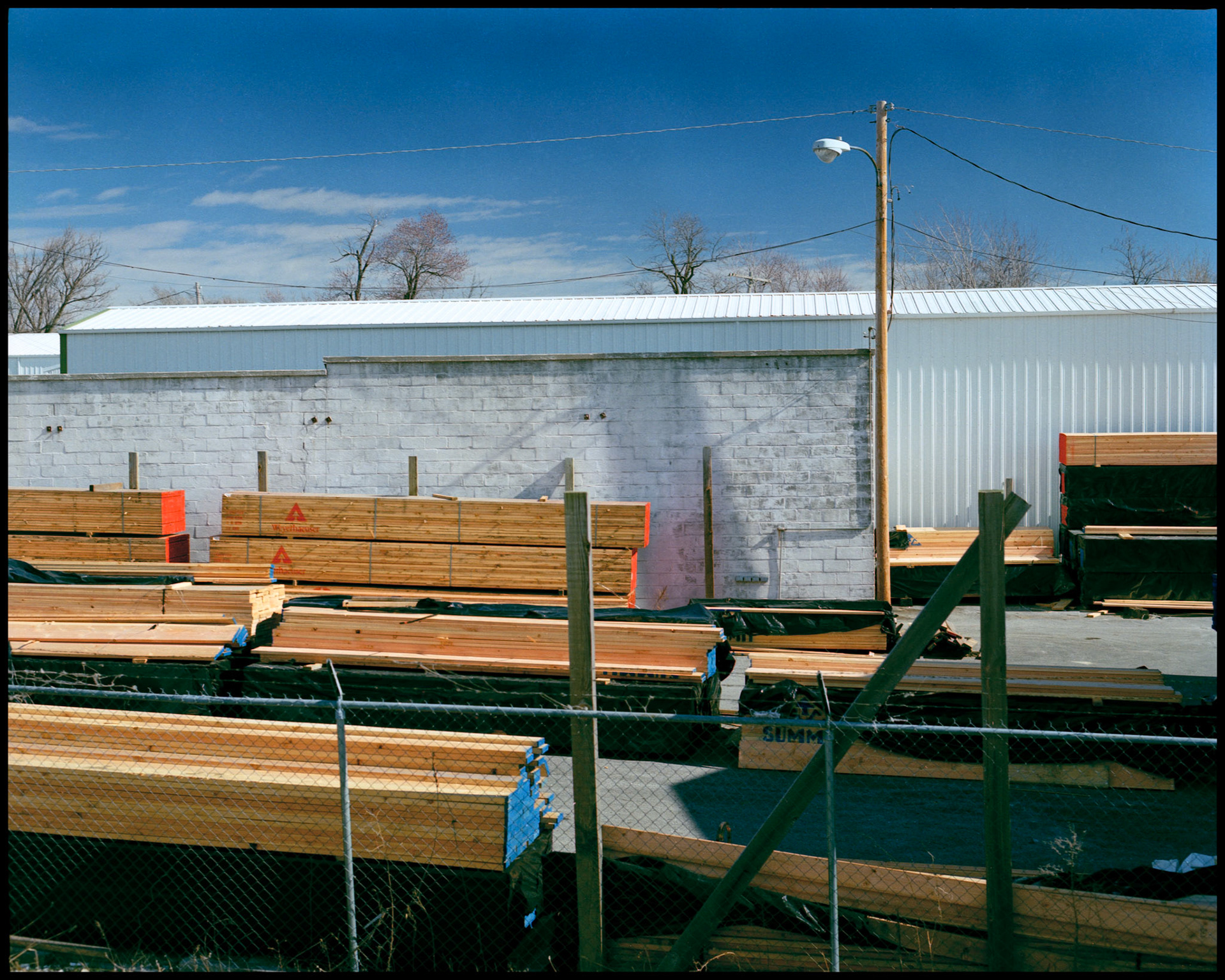 An urban detail view of a lumber yard storage area with colorful stacks of boards in front of a white cement block building with a fence in the foreground. Columbia, Missouri  1990