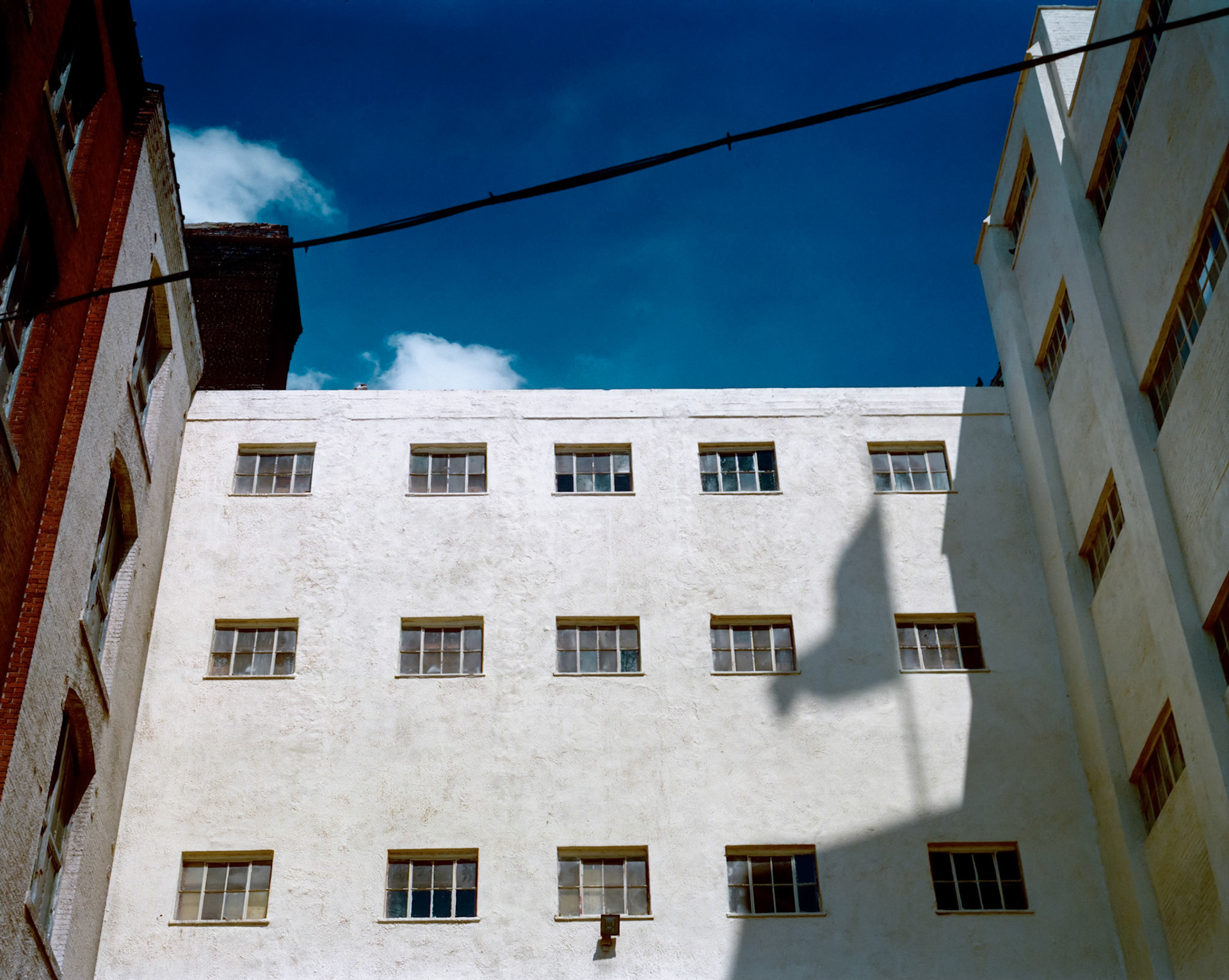 A striking cityscape detail of a brightly lit white stucco wall of a building in the industrial area known as the West Bottoms in Kansas City, Missouri with the shadow of a flowing American flag. 1990