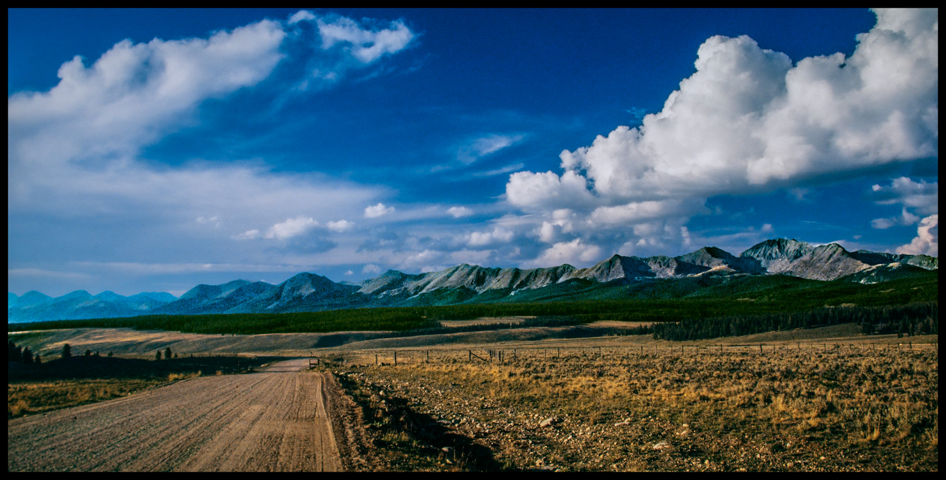 A panorama view of the Collegiate Peaks or Collegiate Range, part of the Sawatch Range in the Rocky Mountains, with billowing clouds against a blue sky as you leave Tin Cup, Colorado on the gravel road to the North. 1985