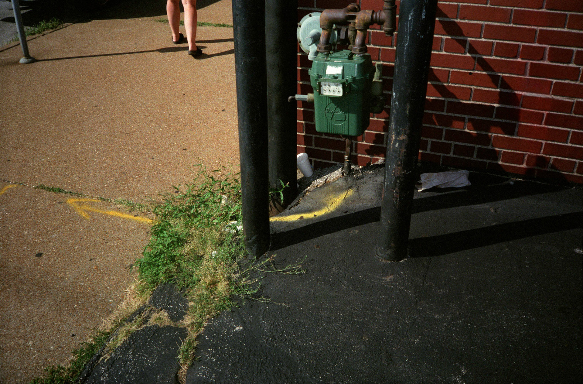 Conceptual abstract detail image of a Natural Gas meter  next to a brick building with safety barrier poles around it and a painted arrow extendning away from it onto a sidewalk. Also on the sidewalk leading out of the frame are a woman's legs. St. Louis, Missouri 1991
