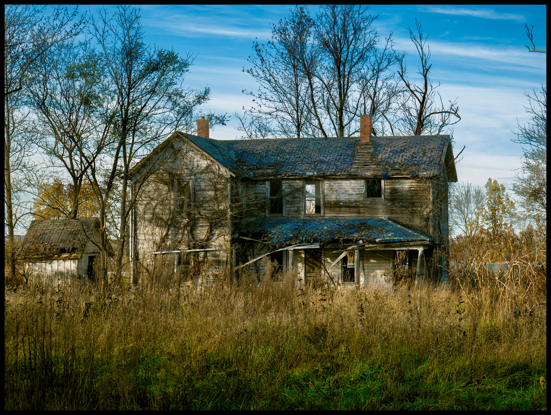 A dilapidated abandoned house with the remains of a blue roof surrounded by desiduous trees and dead weeds lit by the warm light of the late afternoon sun. Near Elmer, Missouri. November 2023