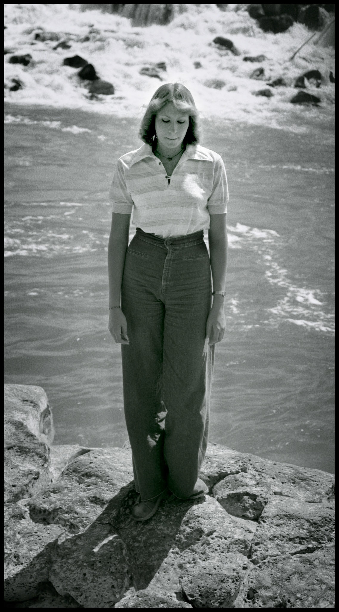 A 1970's vintage image of a young woman deep in thought standing on a rock below Idaho Falls. Idaho Falls, Idaho 1979