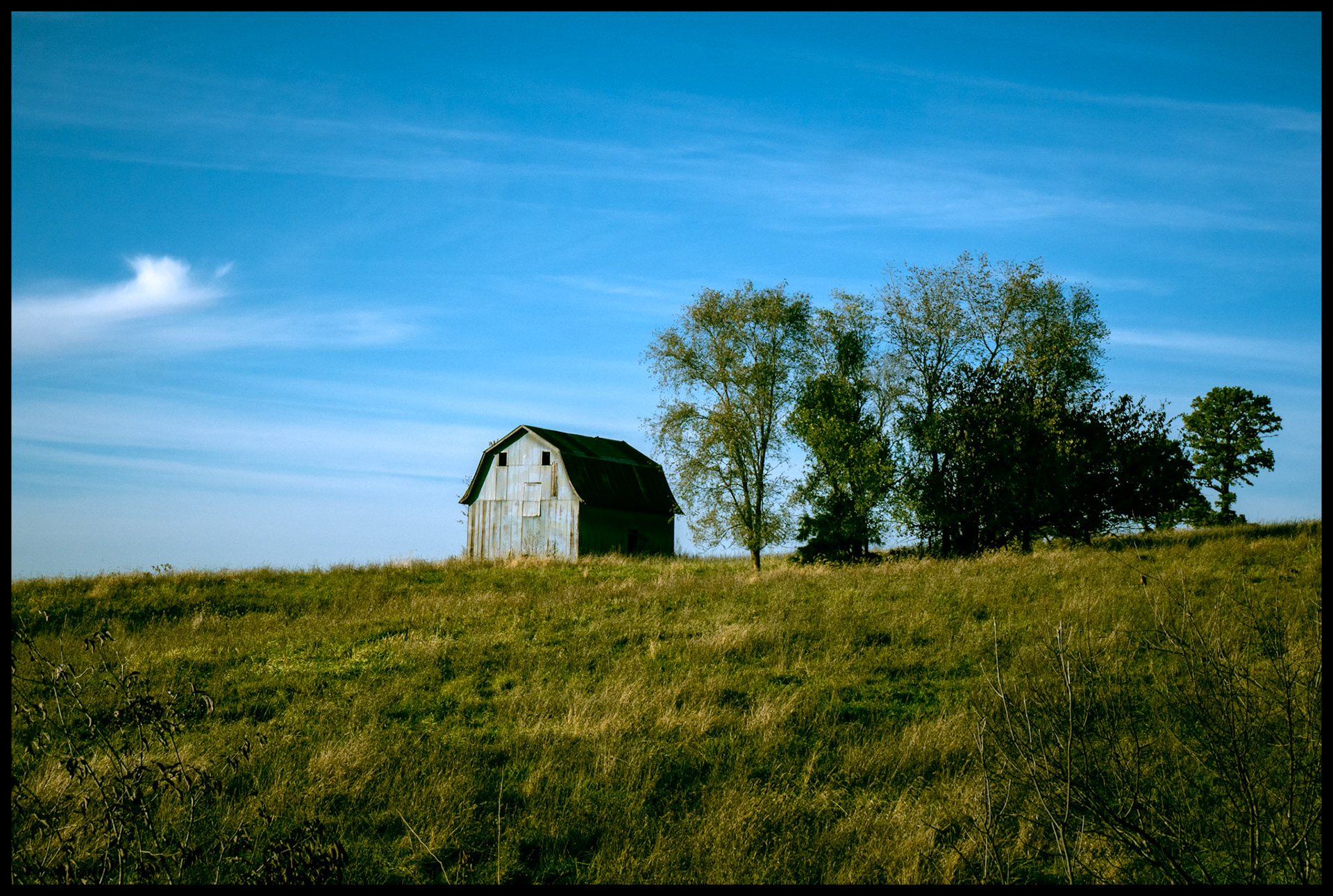 A little barn lit by the late afternoon Autumn sun on a hillside surrounded by trees with a blue sky and wispy cirrus clouds in the background. Near Elmer, Missouri November 2023