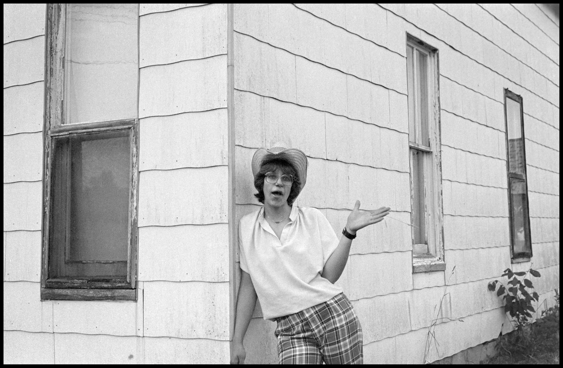 A 1970's gritty vintage image of a young woman in a cowboy hat leaning against a house with a piece of foxtail grass in her hand. Green City, Missouri 1979